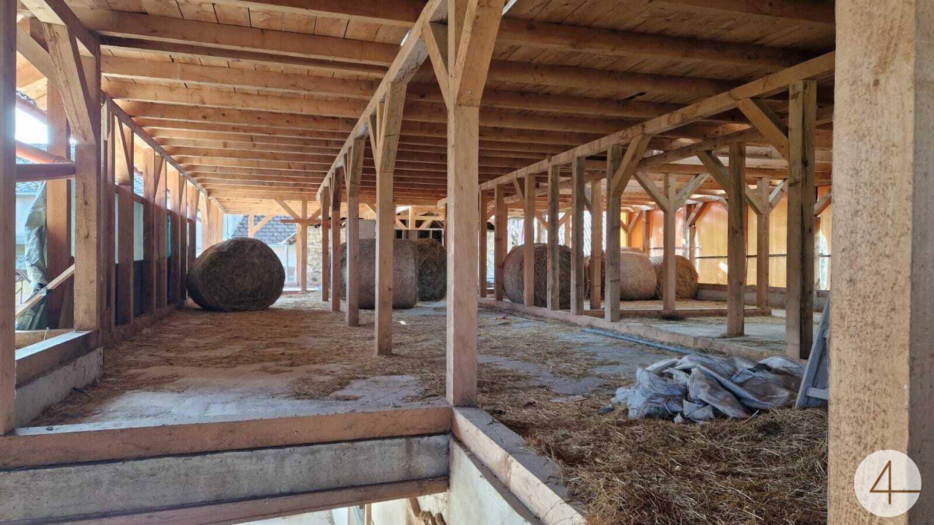 Geräumiger Stall oder Scheunenbereich mit Holzbalken und Heuballen auf dem Boden.