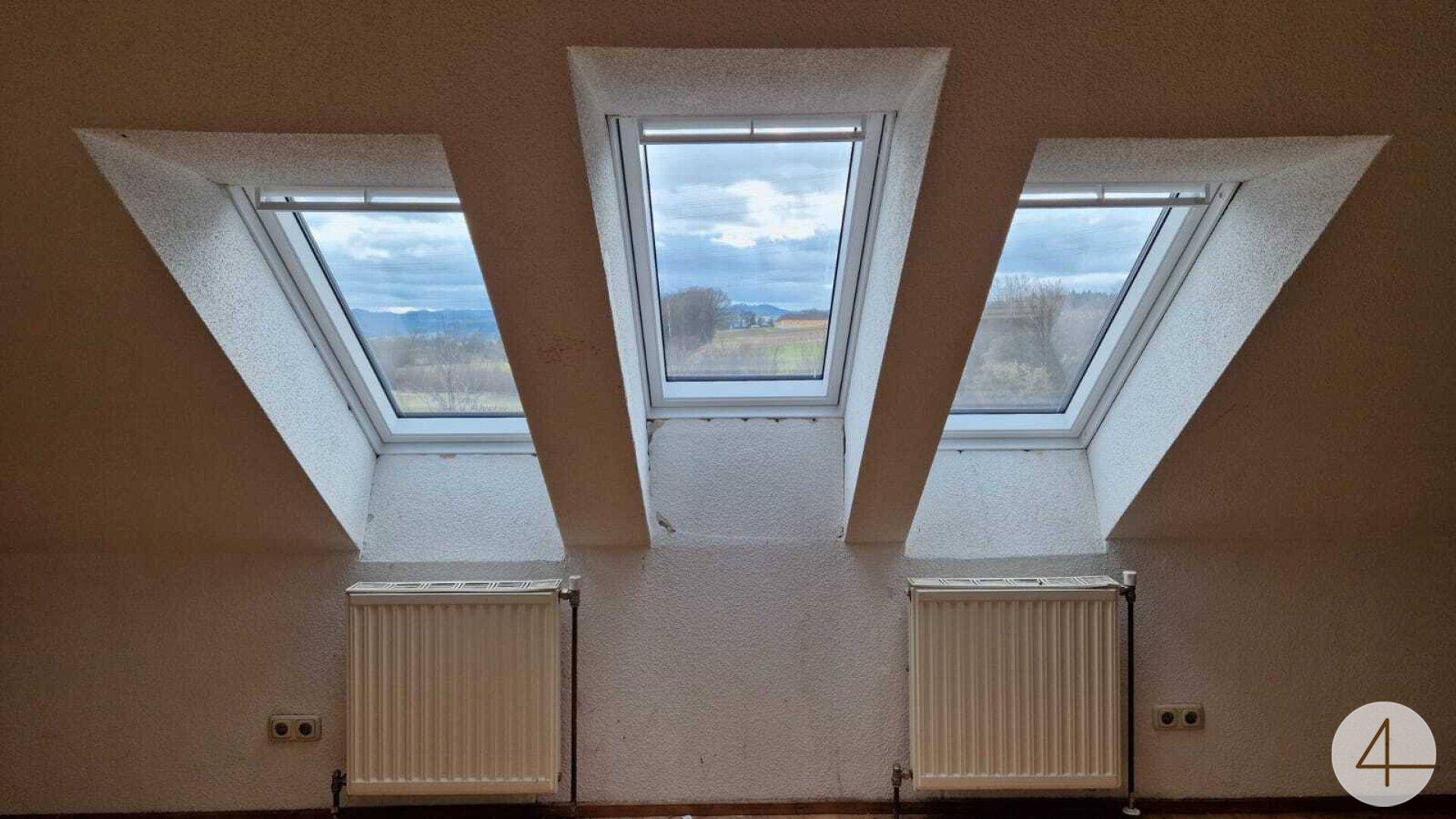 Innenraum mit drei Dachfenstern, die einen weiten Blick auf die Landschaft bieten, und Heizkörpern darunter.