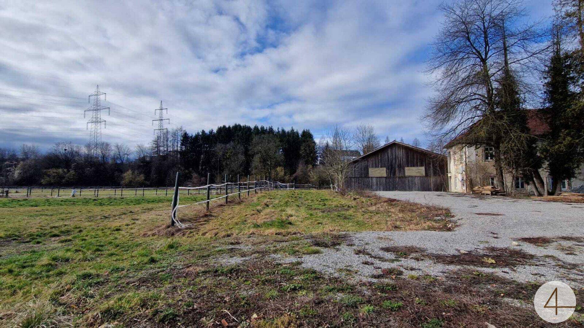 Außenansicht eines Anwesens mit Kiesauffahrt, Holzscheune, altem Steingebäude und eingezäunten Feldern.