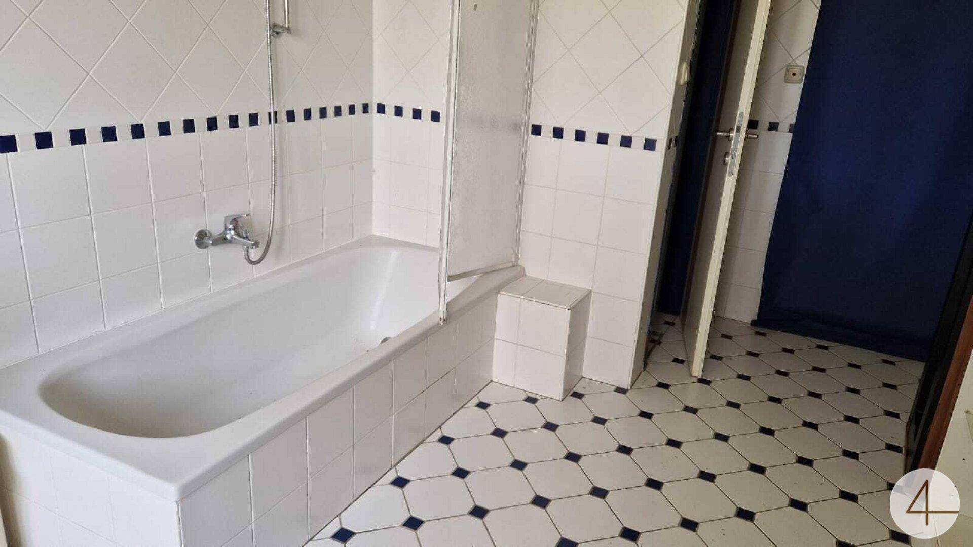 Badezimmer mit Badewanne, weißen Fliesen mit blauem Rand und gemustertem Fliesenboden.