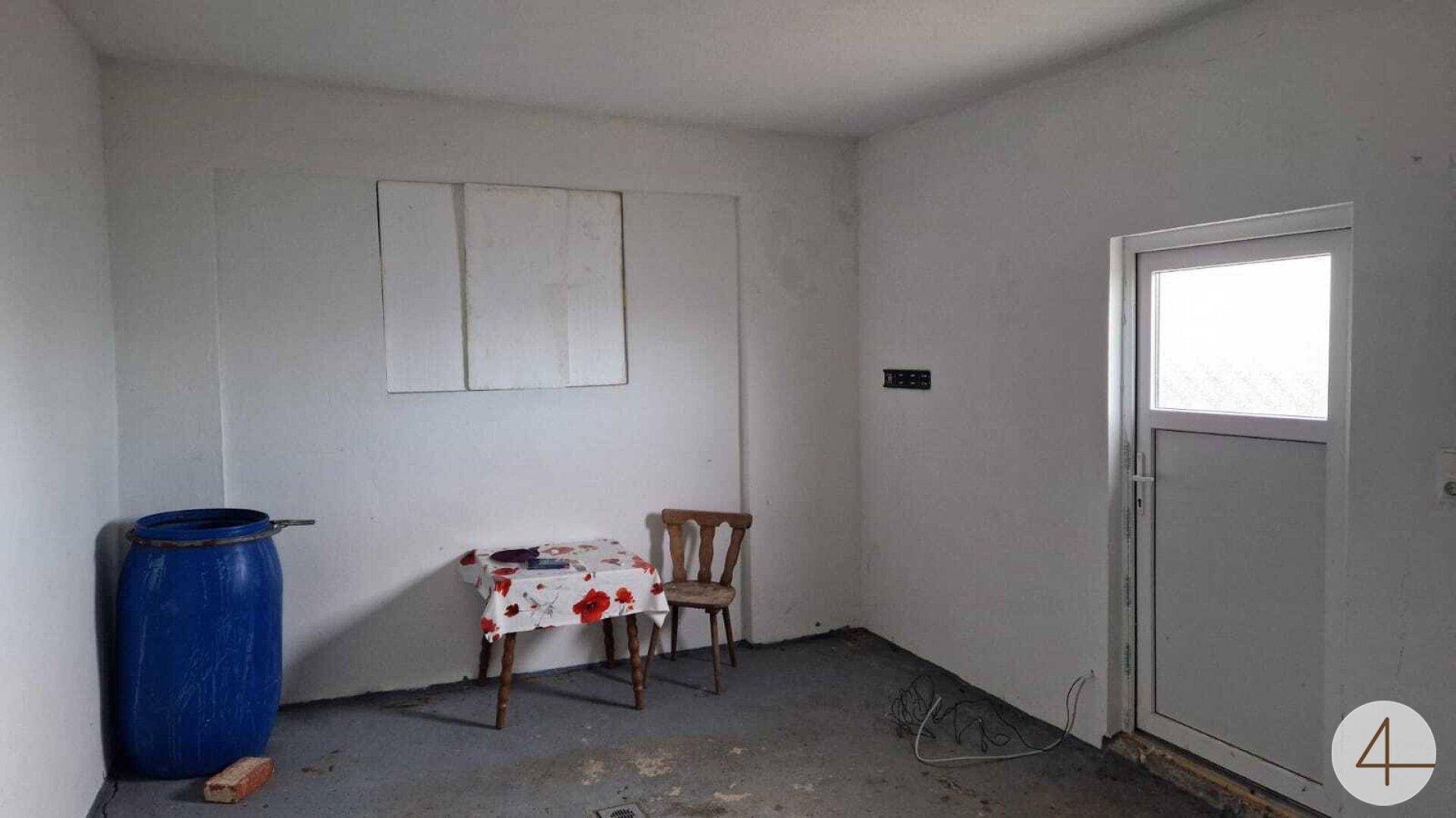 Einfacher Raum mit weißen Wänden, Betonboden, einem Tisch und einer Tür mit Fenster, möglicherweise ein Abstellraum.