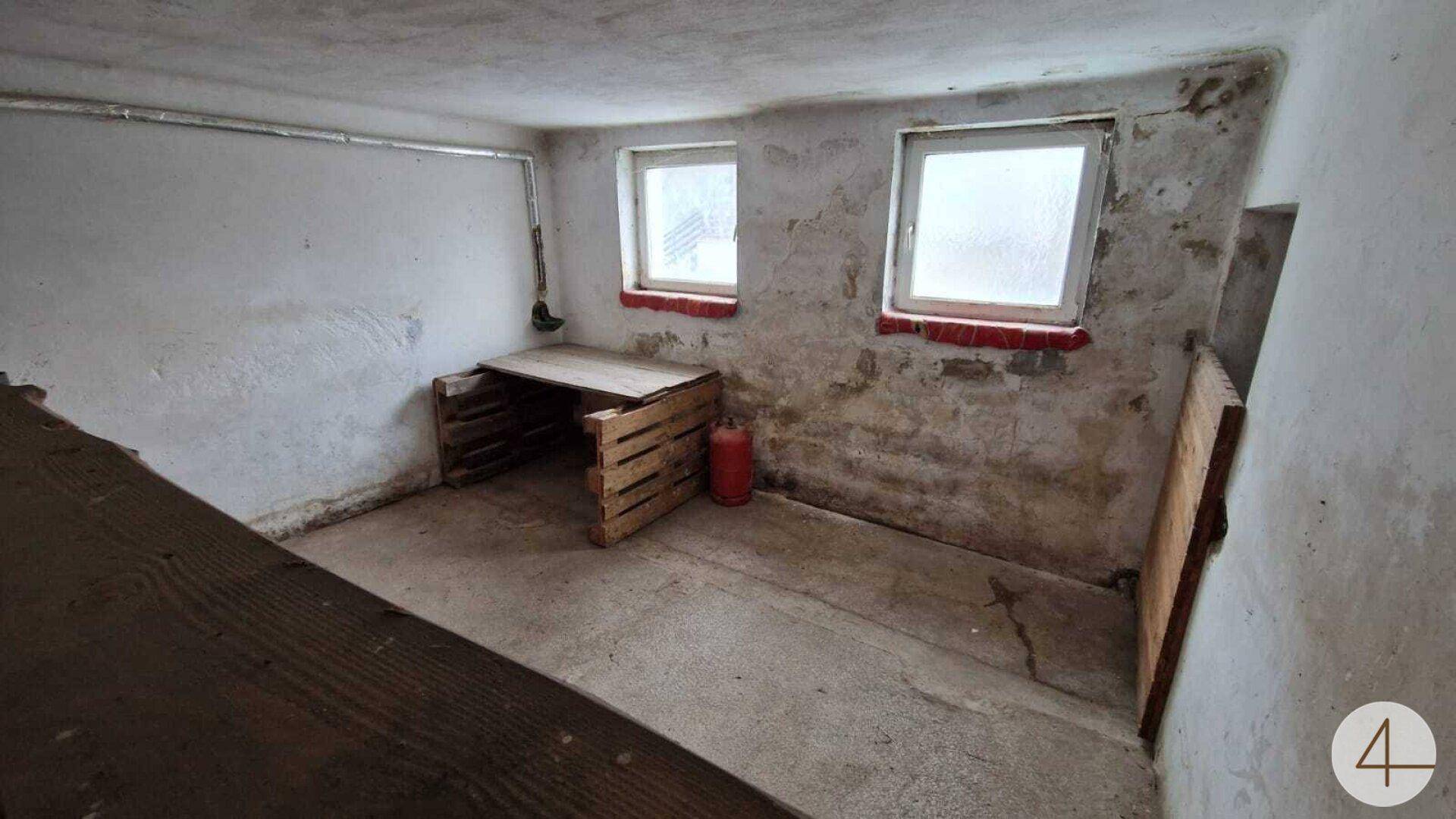 Rustikaler Werkstattraum mit zwei Fenstern, einer einfachen Holzwerkbank und einem Feuerlöscher an der Wand.