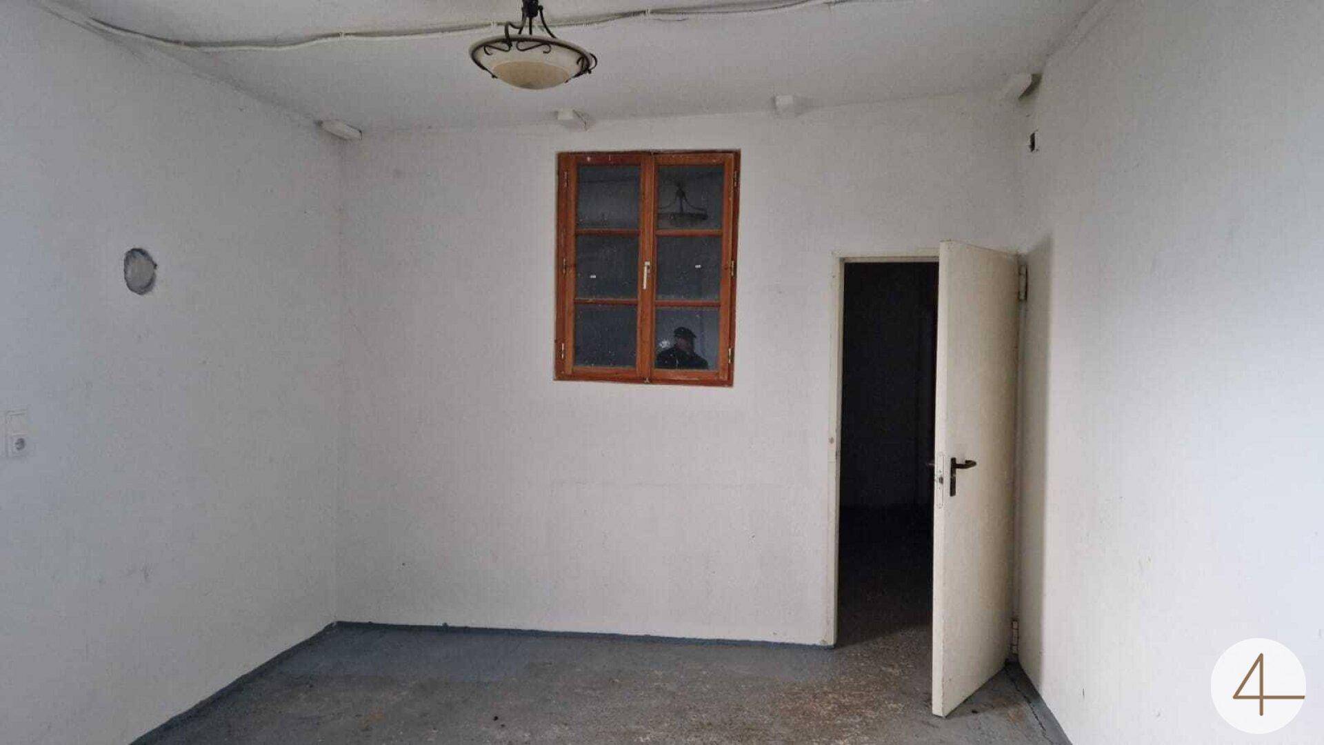Leerer Raum mit einfachen weißen Wänden, einem Holzfenster und einer geöffneten Tür.