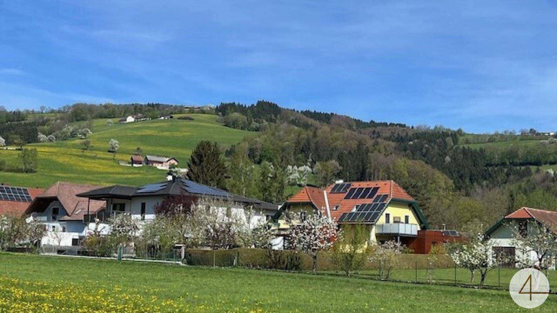 Ländliche Häuser mit Solaranlagen auf den Dächern, umgeben von grünen Hügeln und blühenden Bäumen.