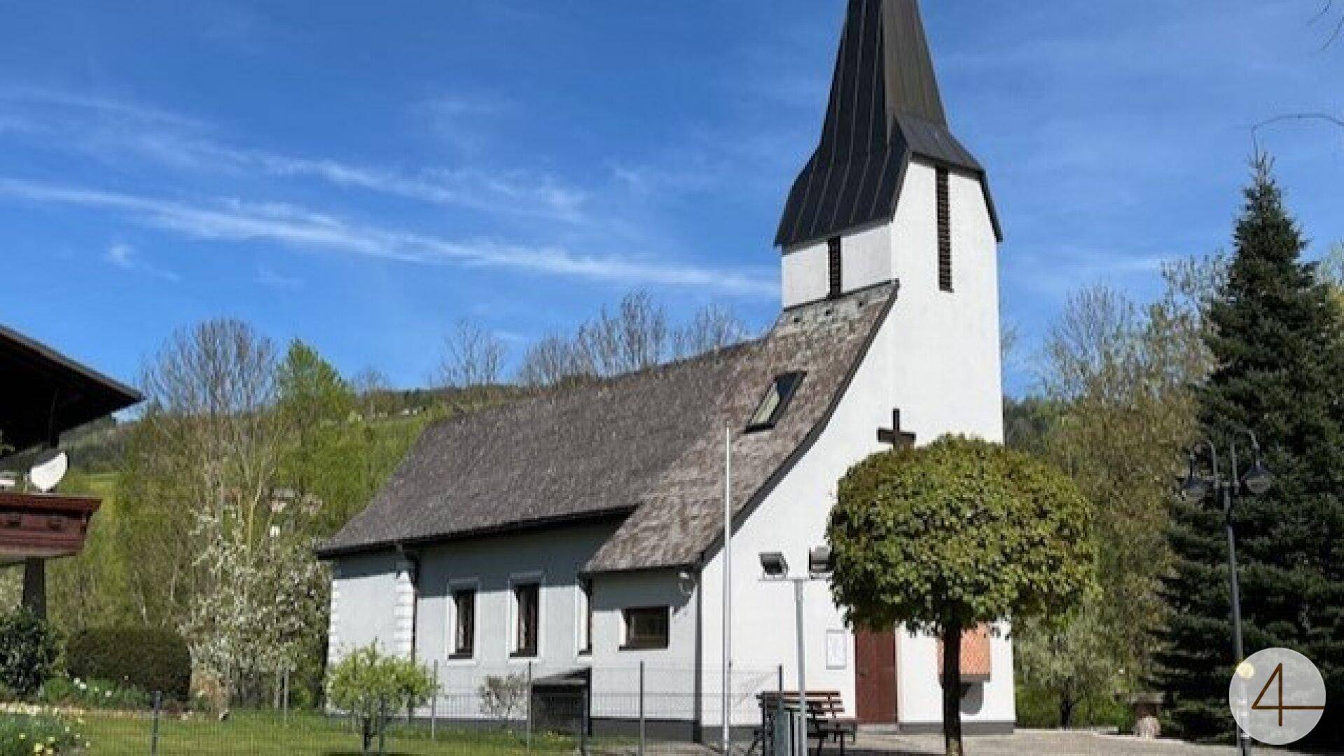 Kleine weiße Kirche mit hohem, spitzem Turm in einer idyllischen ländlichen Umgebung.