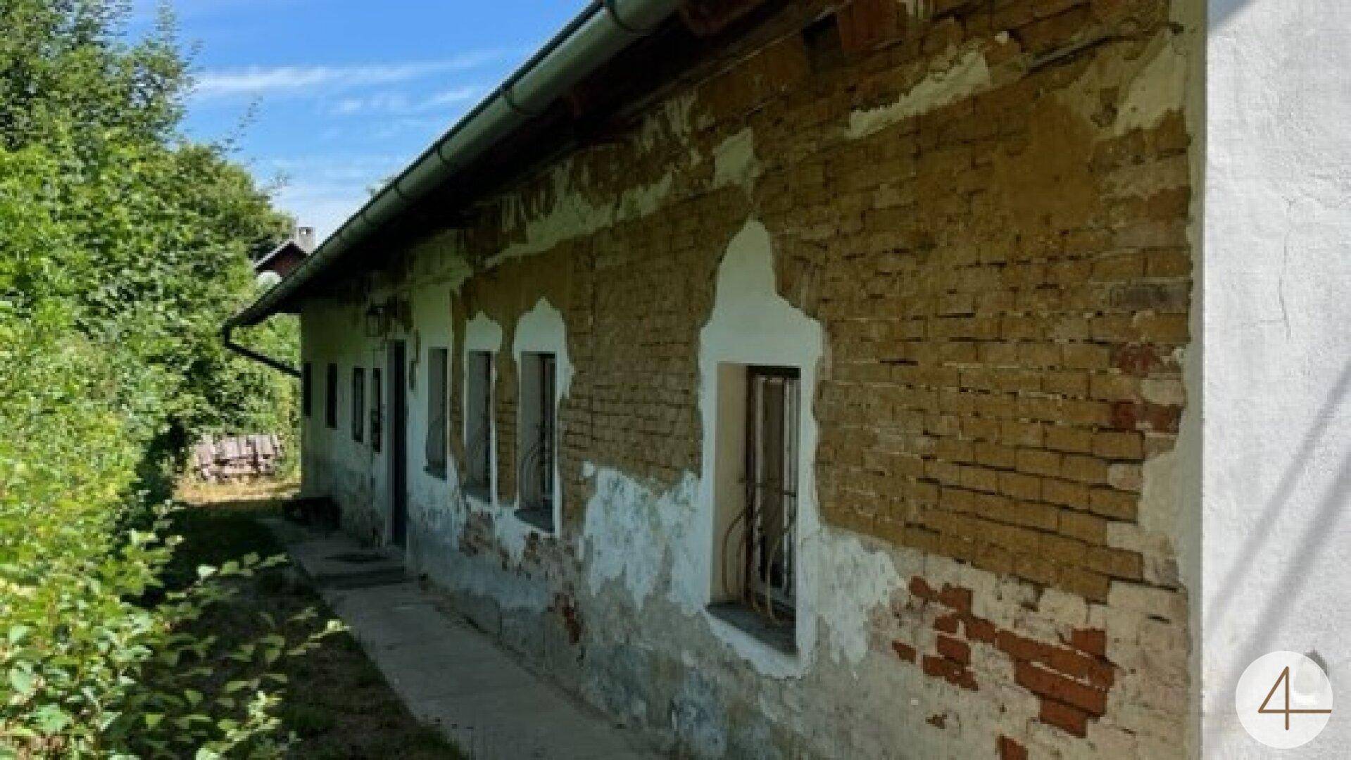 Seitenfassade eines alten Hauses mit abblätterndem Putz, der das Mauerwerk freilegt, und vergitterten Fenstern.