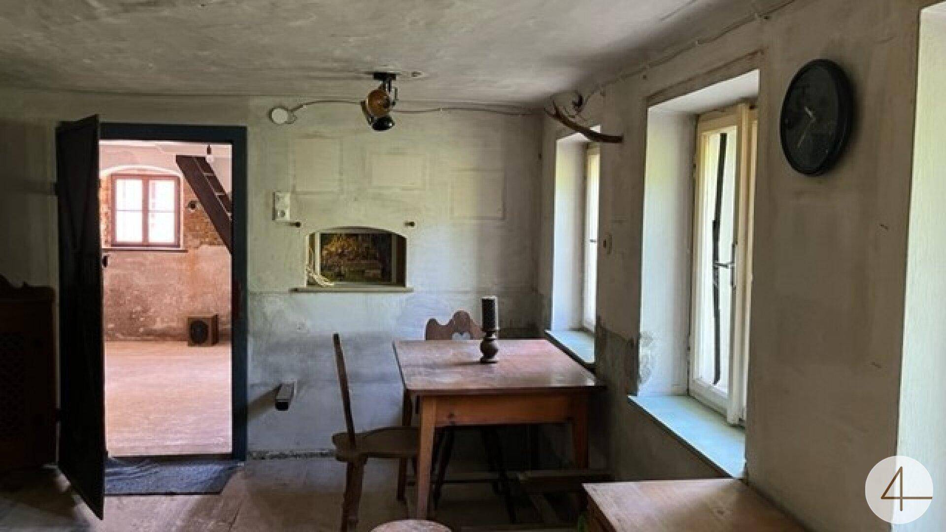 Einfacher Wohnraum mit Holztisch, Stühlen und einem alten Einbauofen in der Wand, angrenzend an einen weiteren Raum.