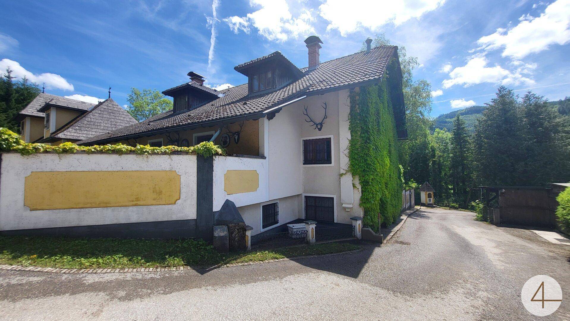 Historisches Gebäude mit gelber Fassade, umgeben von üppigem Grün und Bäumen in ländlicher Umgebung.