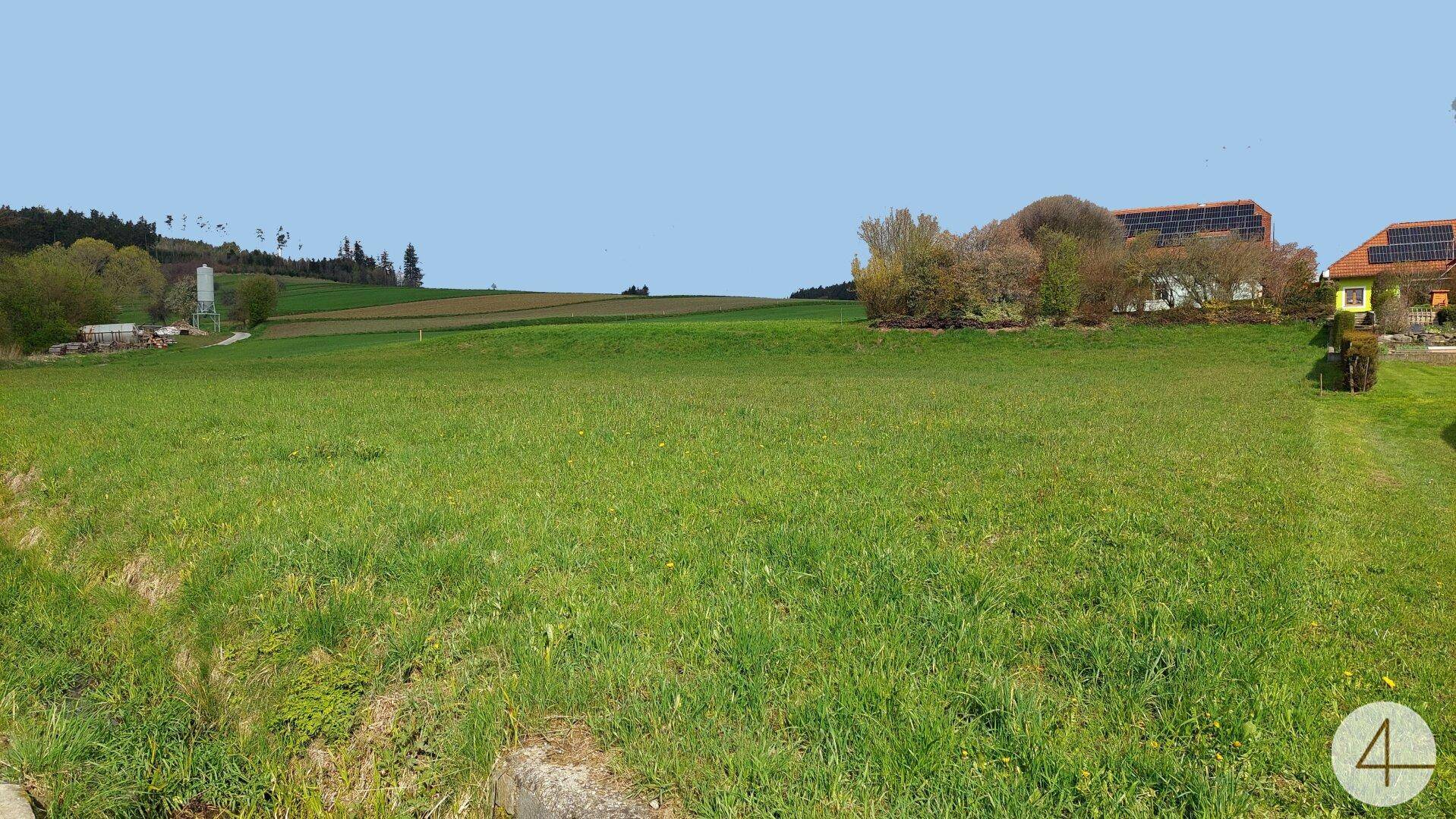 Offene Grünfläche mit Häusern, die Solarpaneele auf den Dächern tragen, und Bäumen am Horizont.