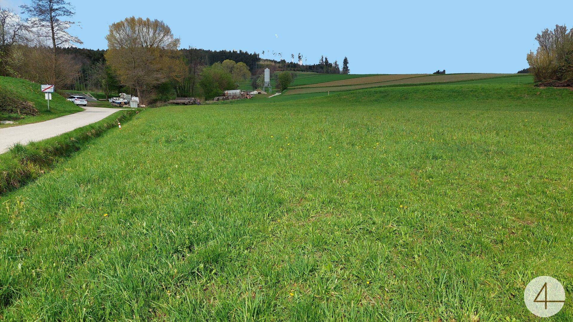 Weite grüne Landschaft mit einer Landstraße und entfernten Gebäuden unter einem weiten blauen Himmel.