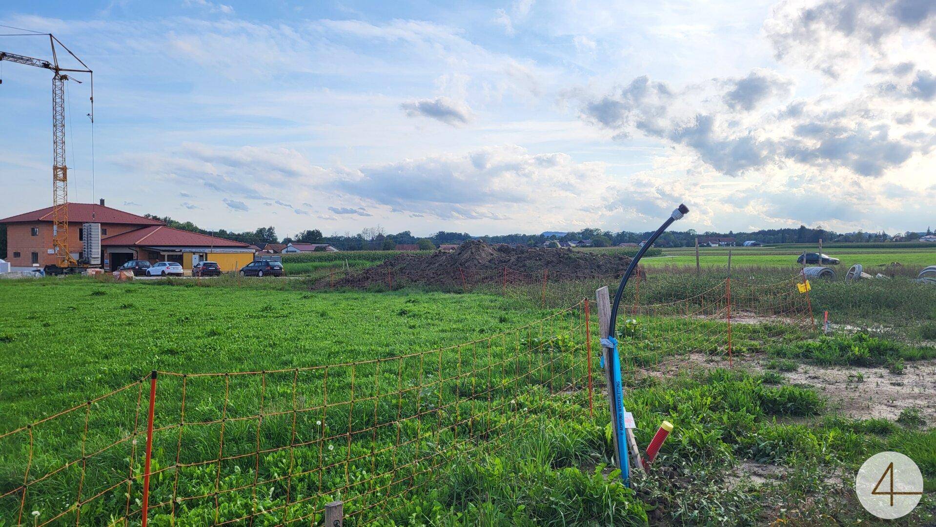 Baustelle mit Erdhaufen und sichtbaren Leitungen im Vordergrund, umgeben von grünen Feldern und einem Gebäude.