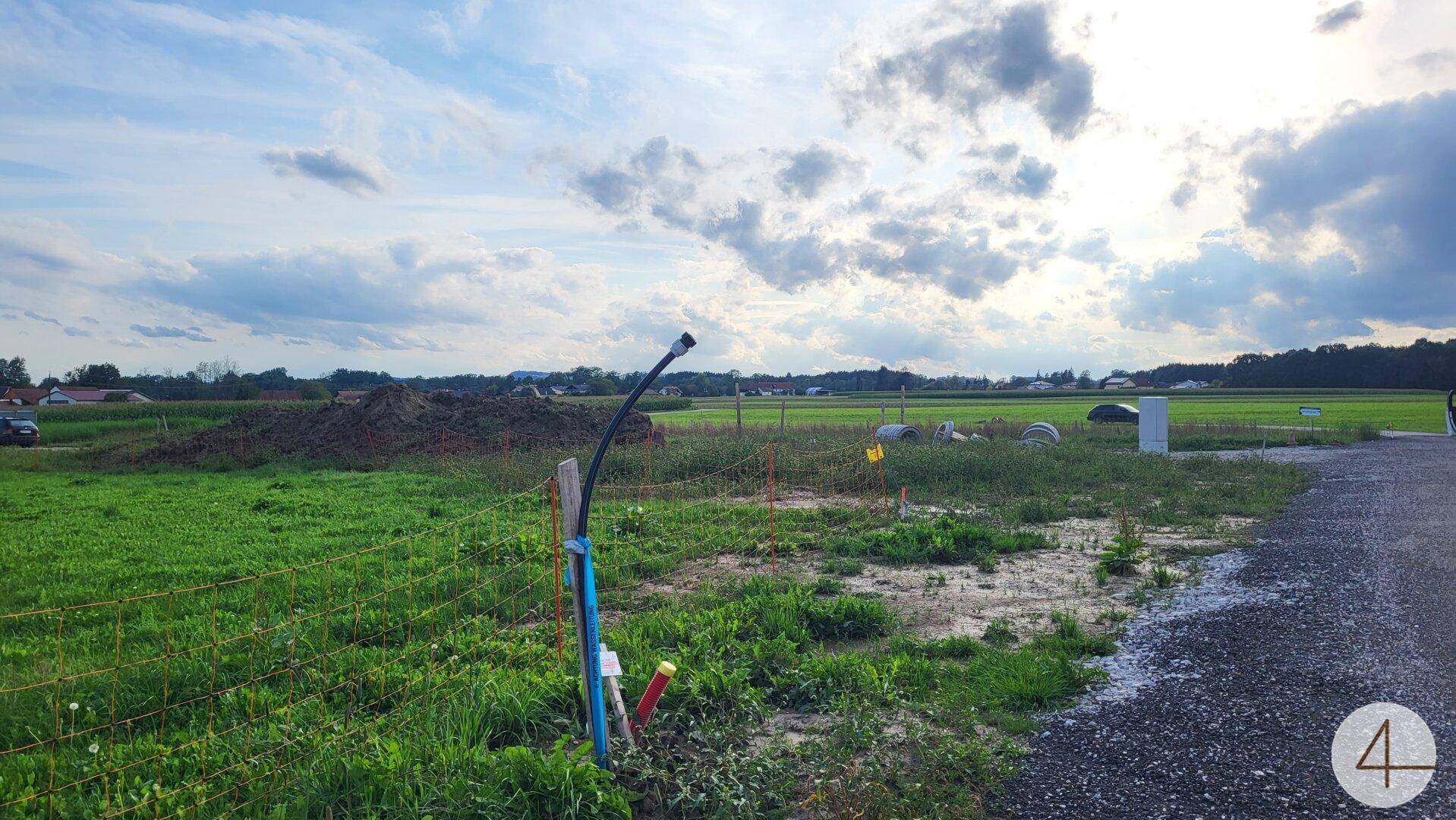Ländliche Baustelle mit Erdhaufen, sichtbaren Versorgungsleitungen und einem Feld unter bewölktem Himmel.
