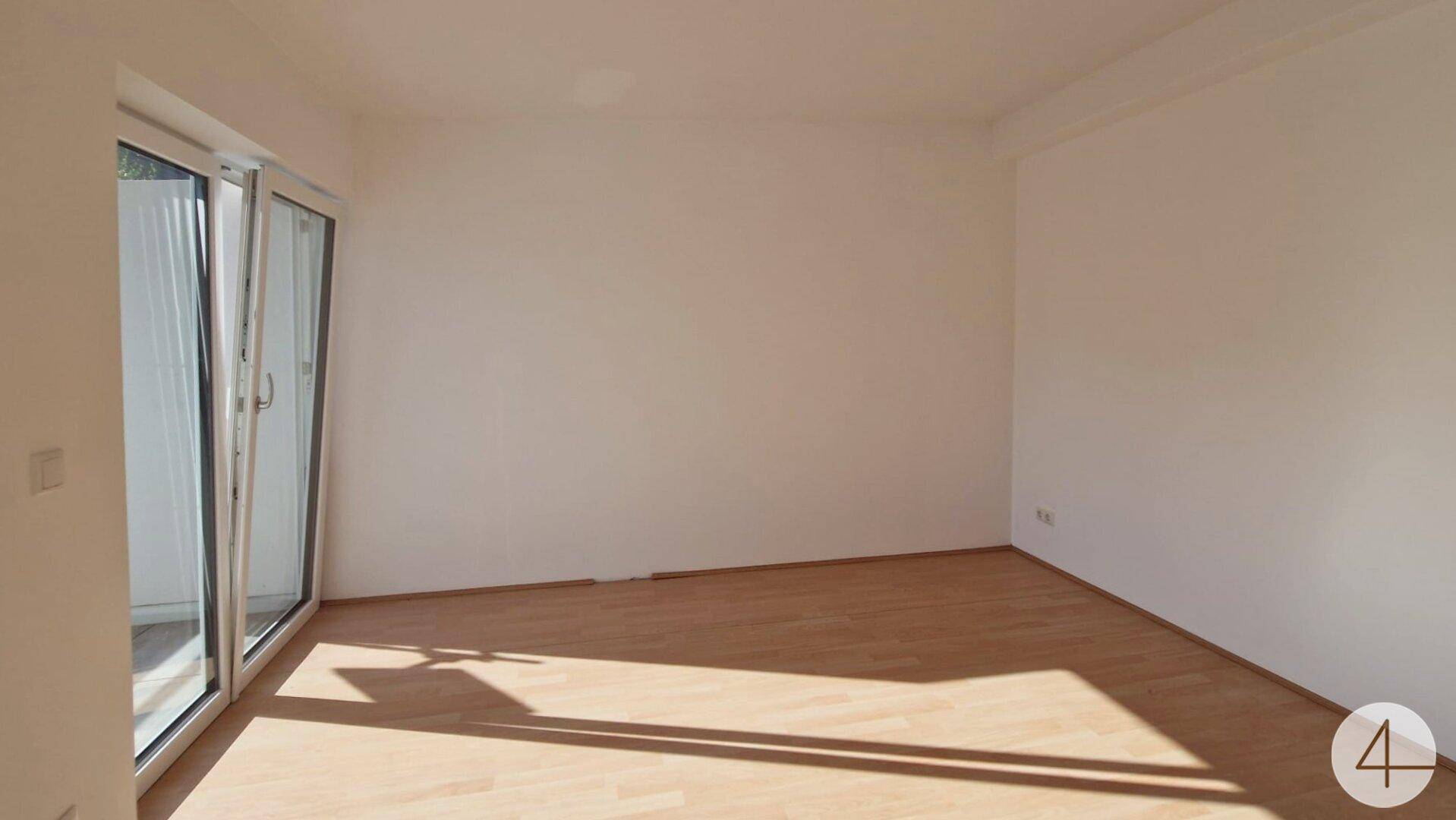 Leeres, helles Zimmer mit Holzboden und Zugang zu einem Balkon, sonnendurchflutet.