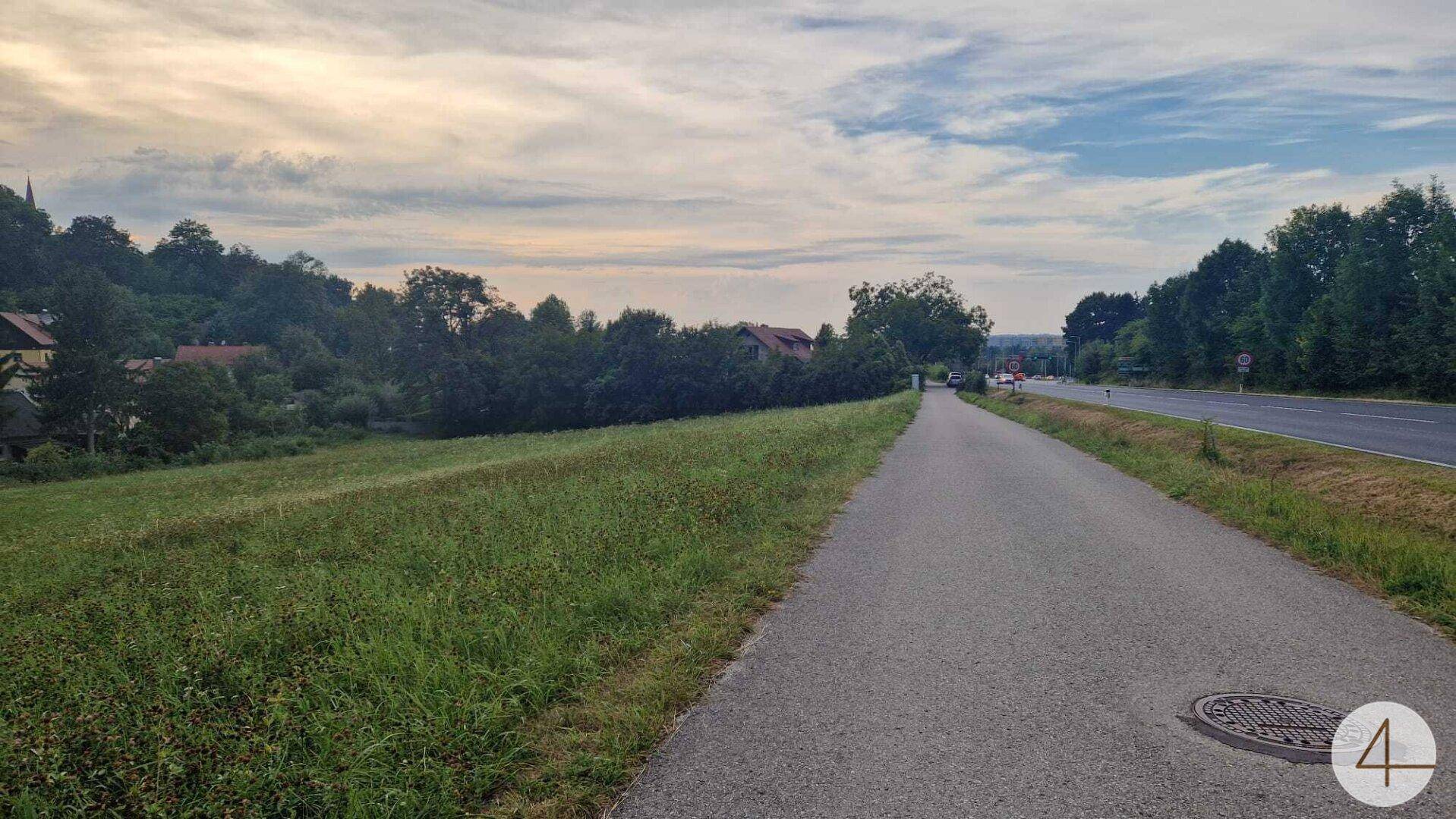 Asphaltierter Weg neben einem grünen Feld mit ländlicher Bebauung und weitem Blick.