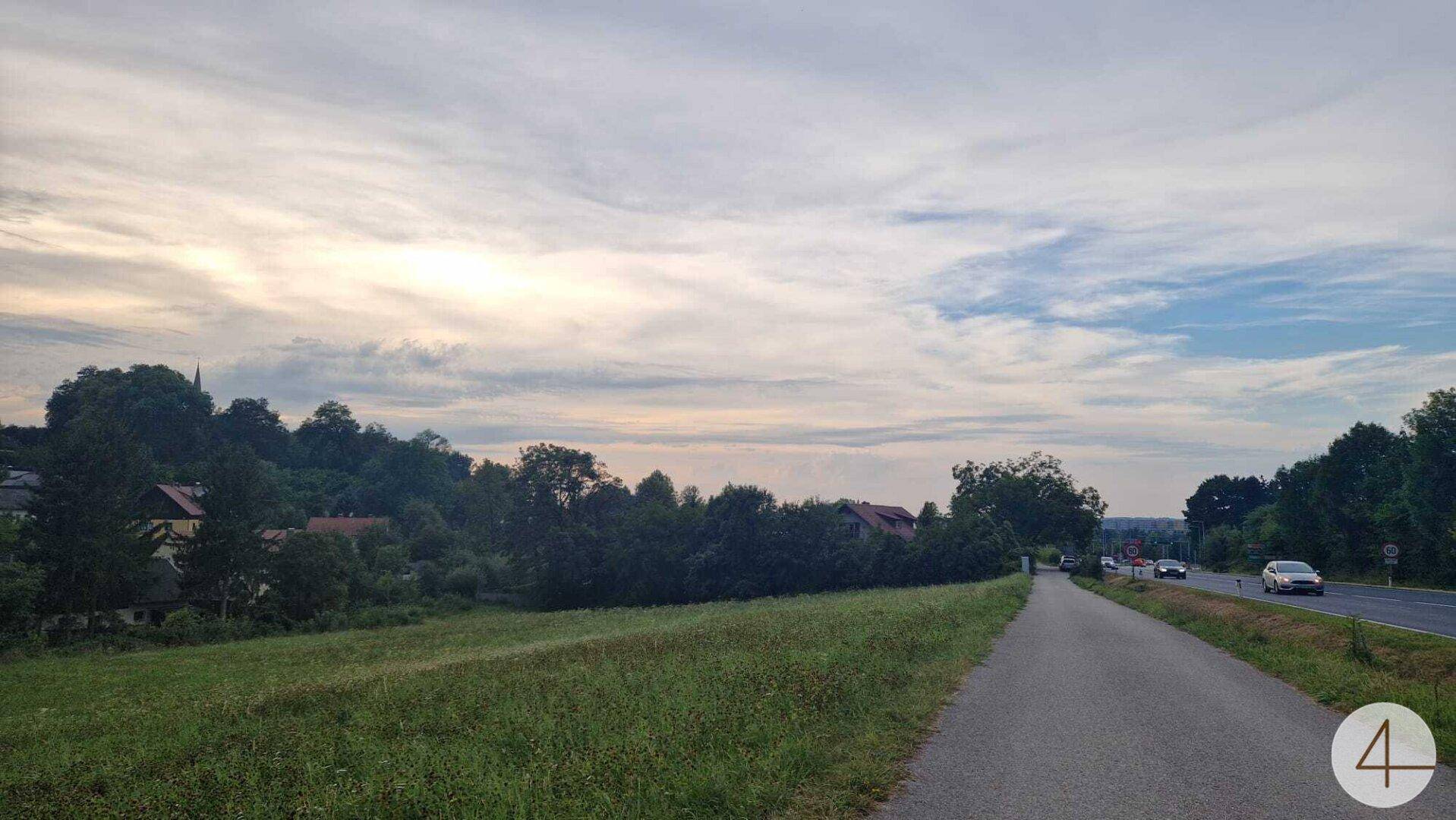 Asphaltierter Weg neben einem grünen Feld mit ländlicher Bebauung und weitem Blick.