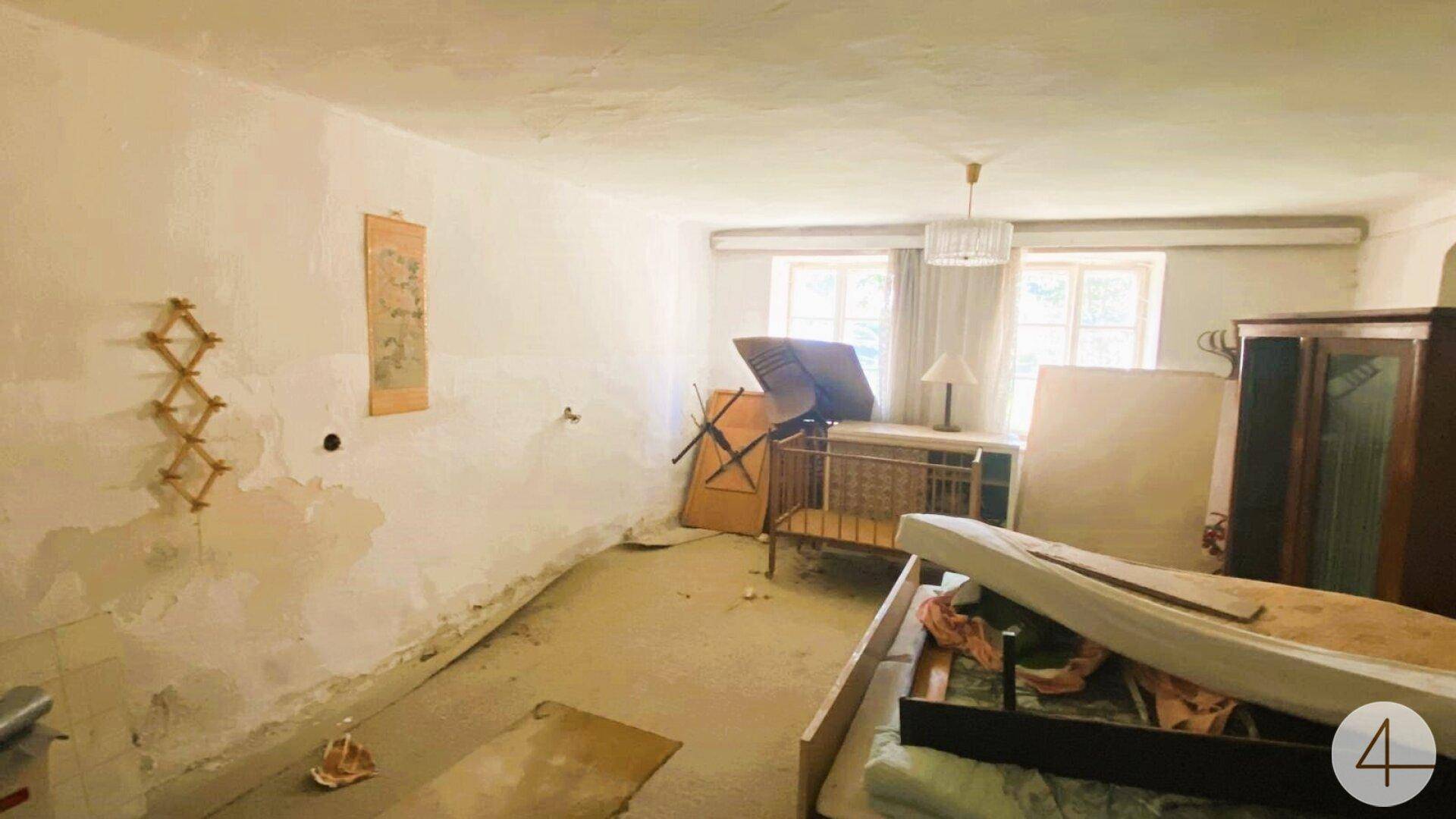 Schlafzimmer mit alten Möbeln, abblätternden Wänden und zwei Fenstern für natürliches Licht, renovierungsbedürftig.