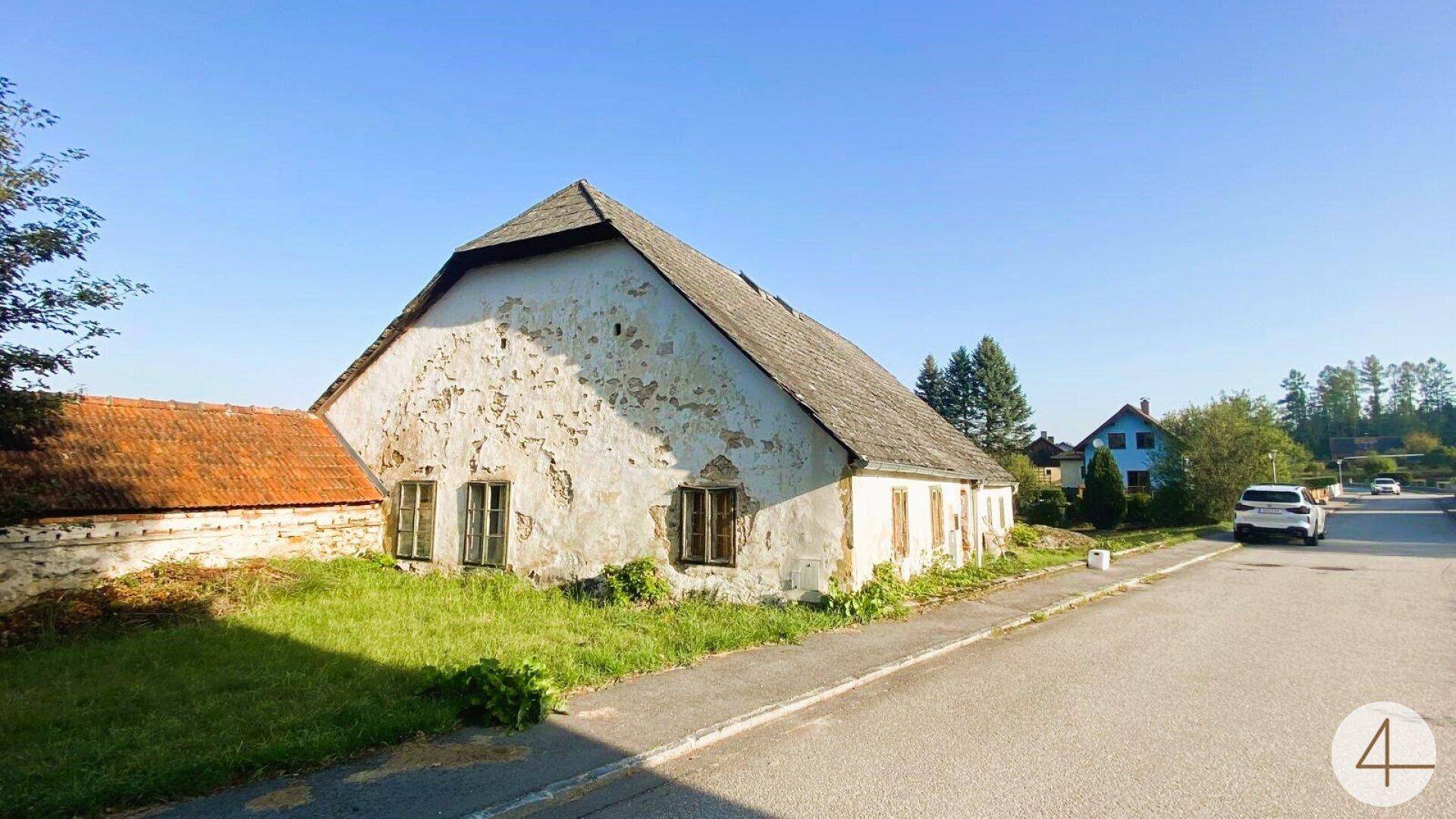 Altes Bauernhaus an einer ländlichen Straße mit abblätterndem Putz und einem kleinen Vorgarten unter blauem Himmel.