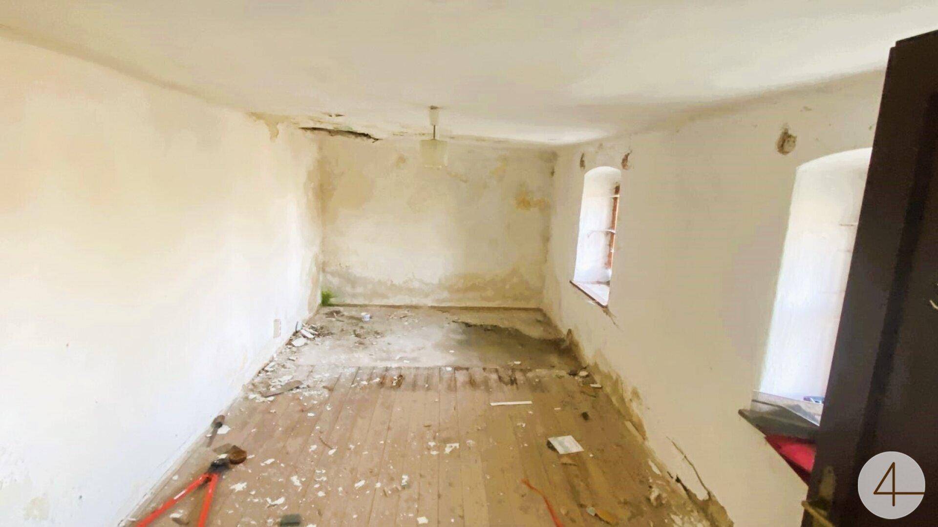 Leerer Raum mit abgenutztem Holzboden, beschädigten Wänden und einem kleinen Fenster, das natürliches Licht hereinlässt.