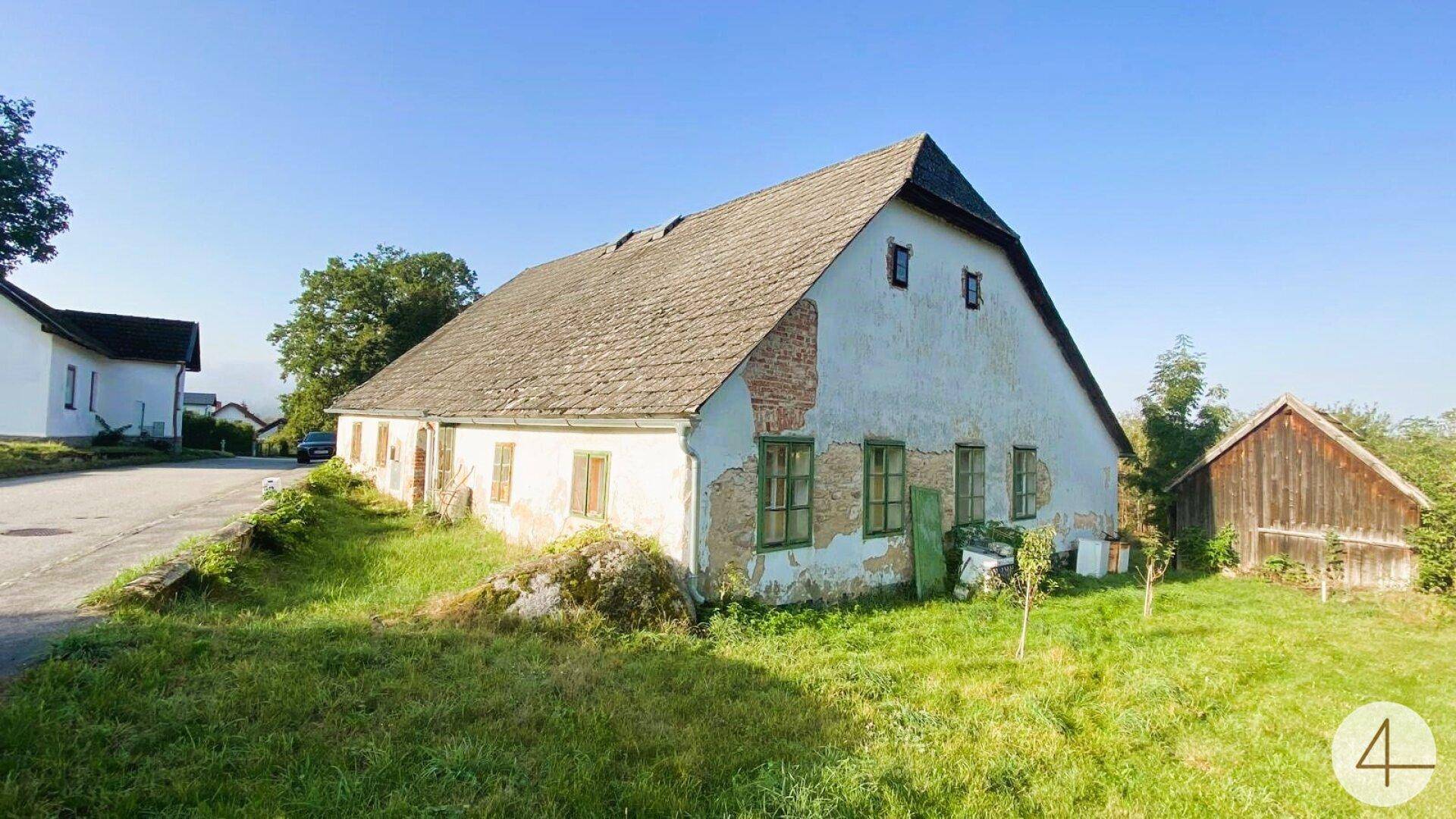 Altes Bauernhaus mit abblätterndem Putz, grünen Fenstern und einem kleinen Holzschuppen daneben an einer Straße.