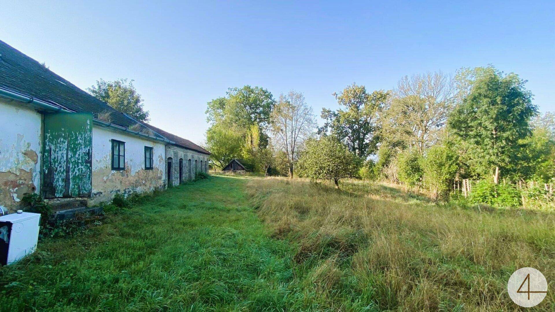 Lange Fassade eines alten Gebäudes mit abblätterndem Putz und grünen Fensterrahmen, umgeben von einer weiten Wiese.