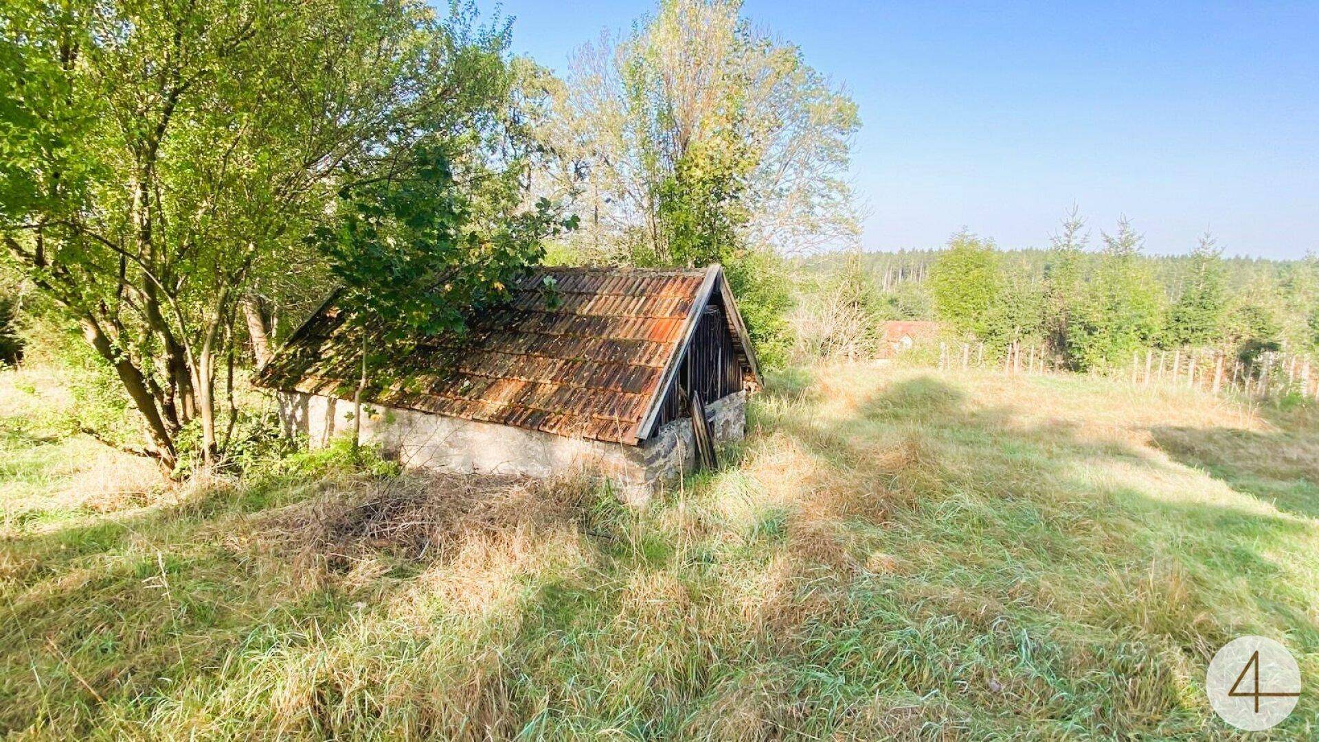 Kleines Nebengebäude mit altem Ziegeldach und Steinmauern in einer sonnigen, ländlichen Umgebung mit viel Grün.