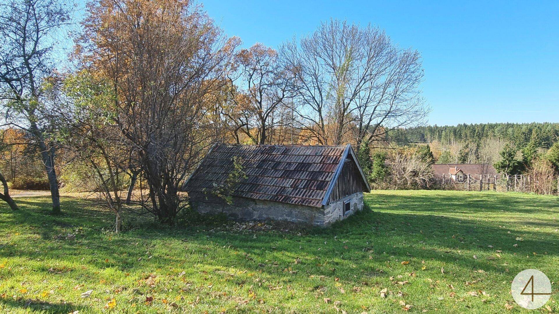 Kleines Nebengebäude mit altem Ziegeldach und Steinmauern, eingebettet in eine grüne Landschaft mit Bäumen.