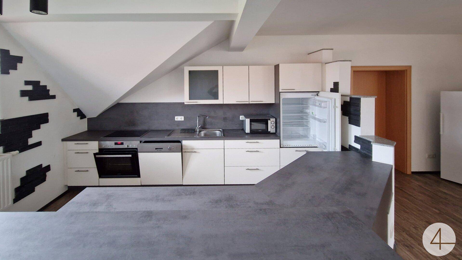 Moderne Einbauküche mit weißen Schränken, grauer Arbeitsplatte und integrierten Geräten unter der Dachschräge.