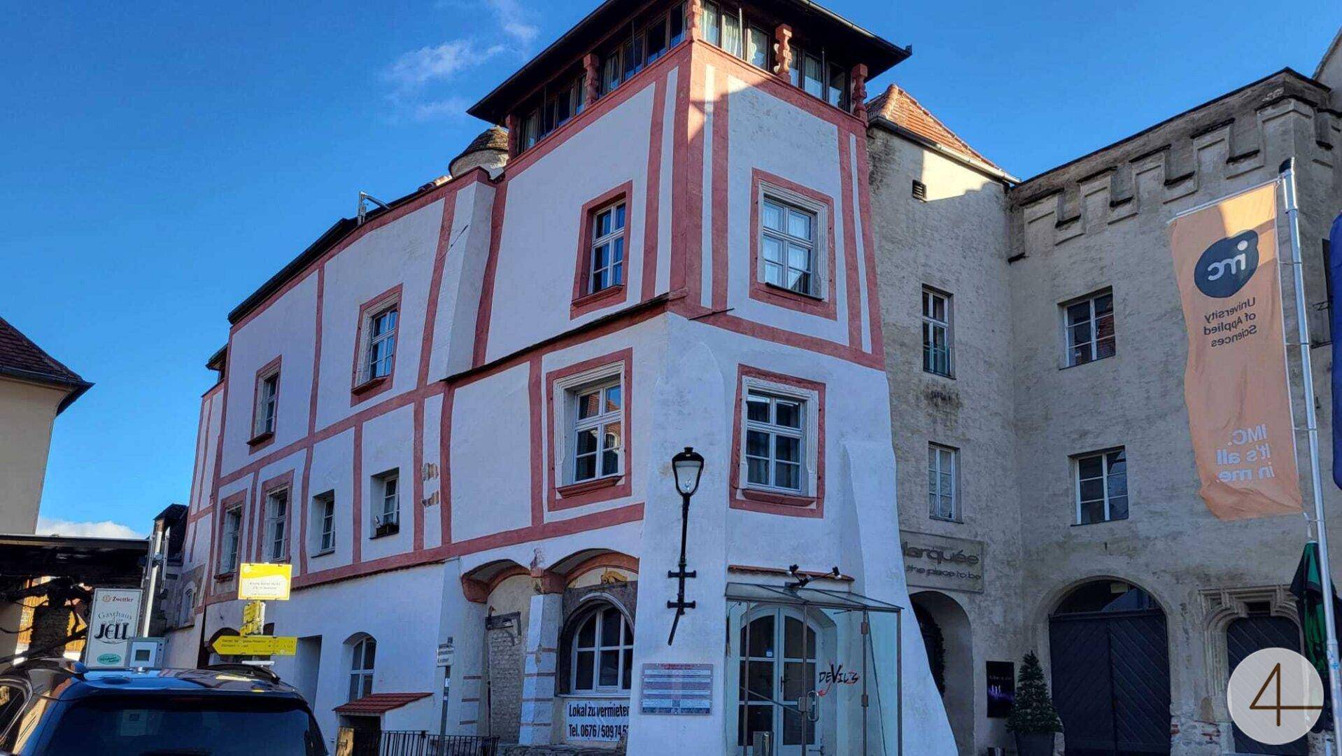 Historisches Gebäude mit markanter Fassade und gepflastertem Vorplatz unter blauem Himmel.