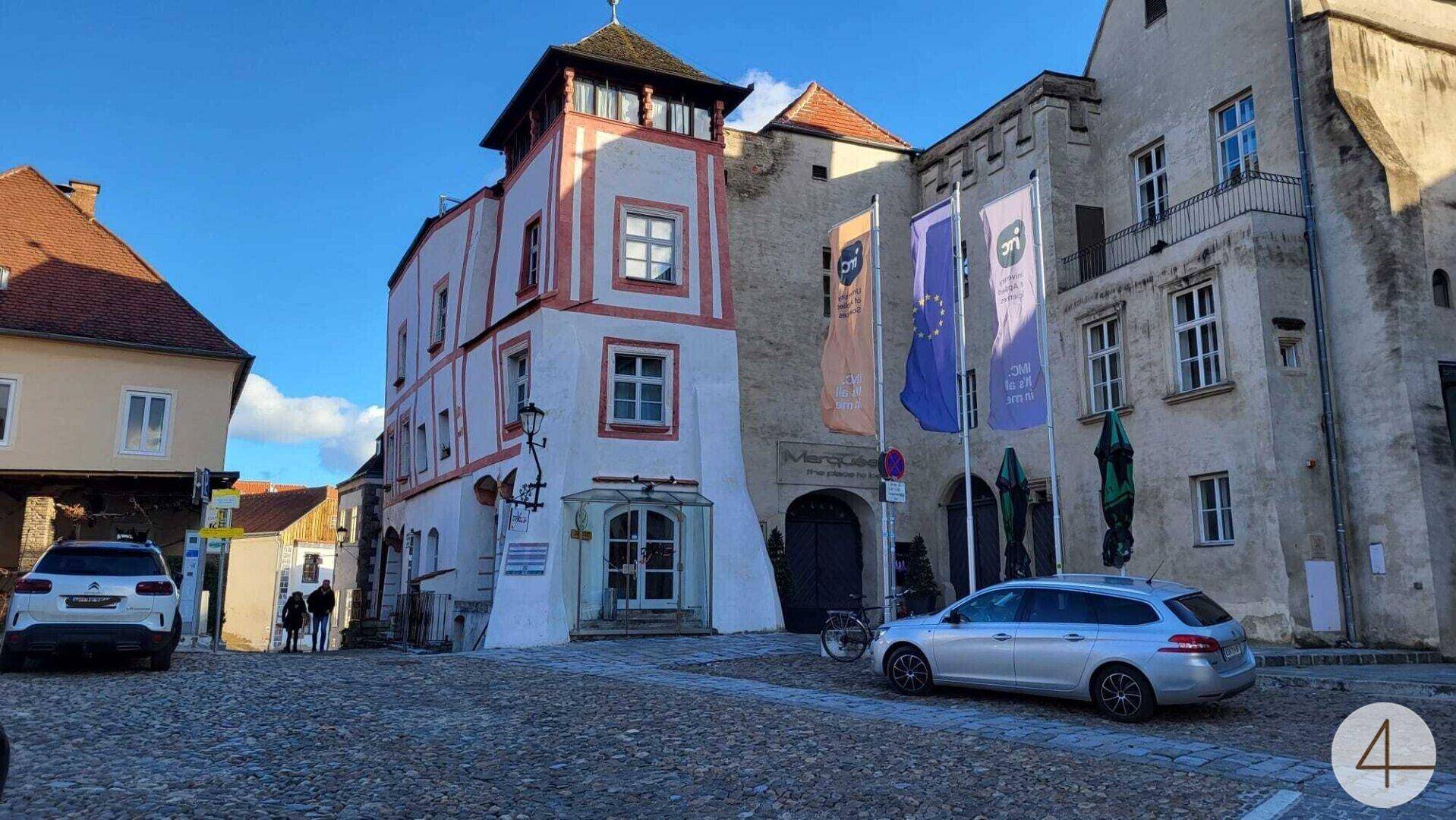 Historisches Gebäude mit markanter Fassade und gepflastertem Vorplatz, umgeben von parkenden Autos.