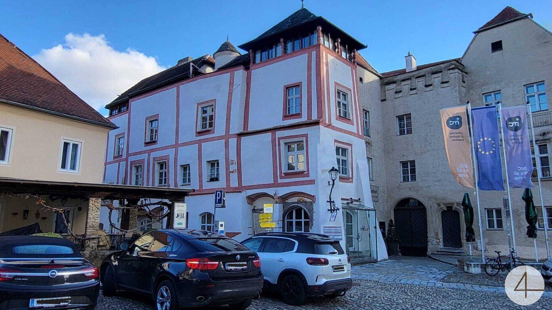 Historisches Gebäude mit markanter Fassade und gepflastertem Vorplatz, umgeben von parkenden Autos.