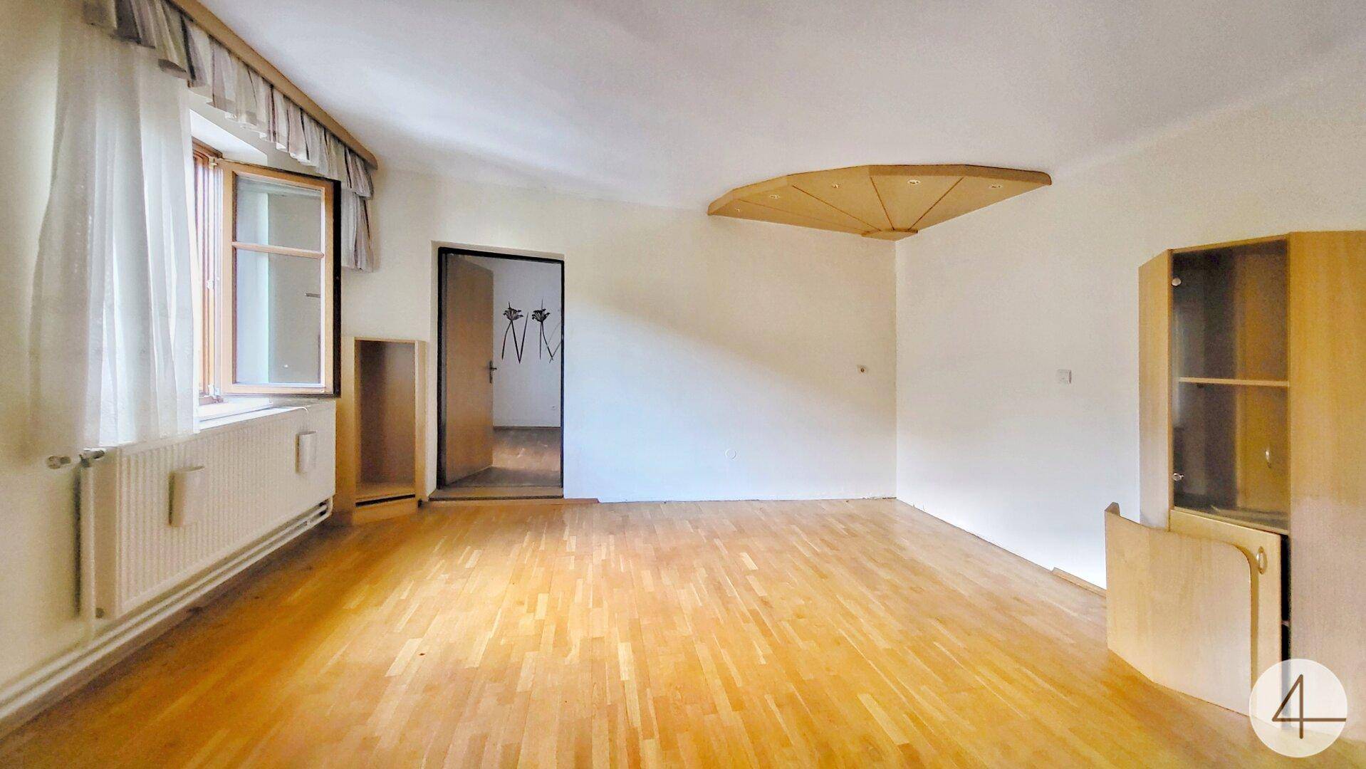 Geräumiges Wohnzimmer mit Parkettboden, großem Fenster und Heizkörper unter der Fensterbank.