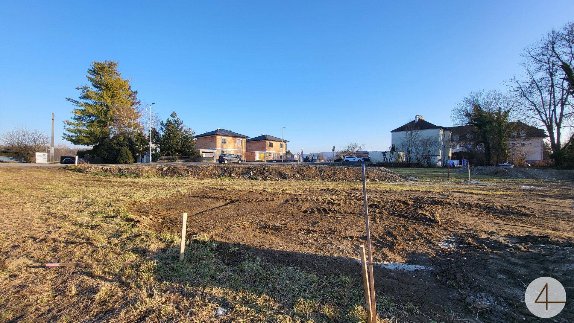 Blick über das unbebaute Grundstück mit vorbereitetem Boden und angrenzenden Wohnhäusern.