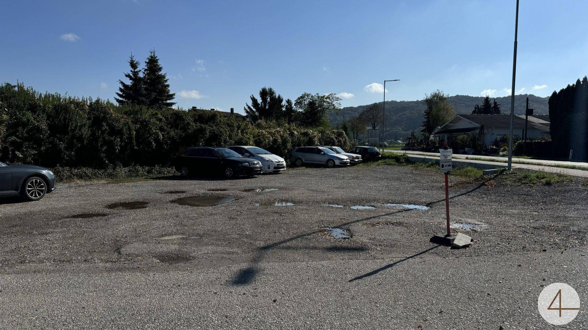 Großer Schotterparkplatz mit mehreren geparkten Autos und grünen Hügeln im Hintergrund unter blauem Himmel.