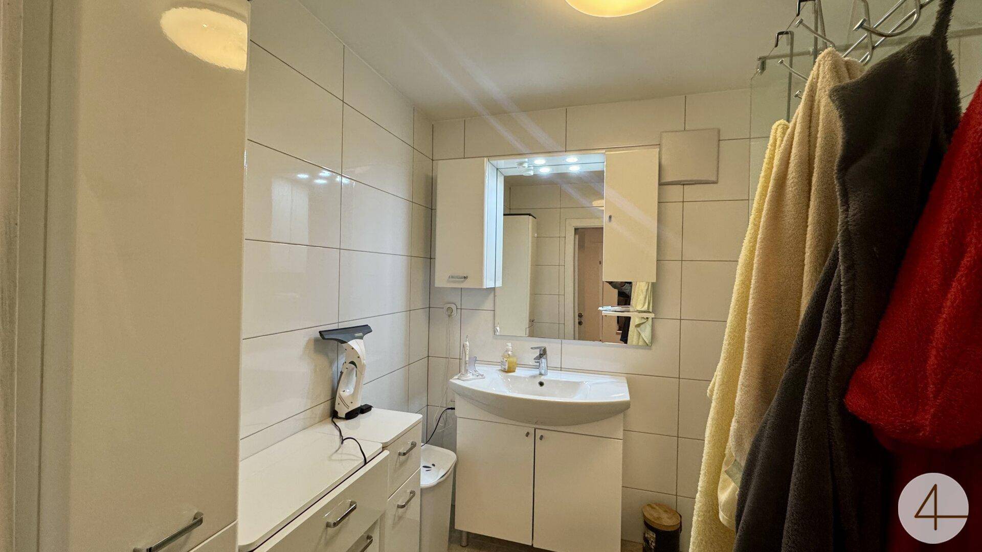 Helles Badezimmer mit weißen Fliesen, Waschbecken, Spiegelschrank und praktischen Stauschränken.