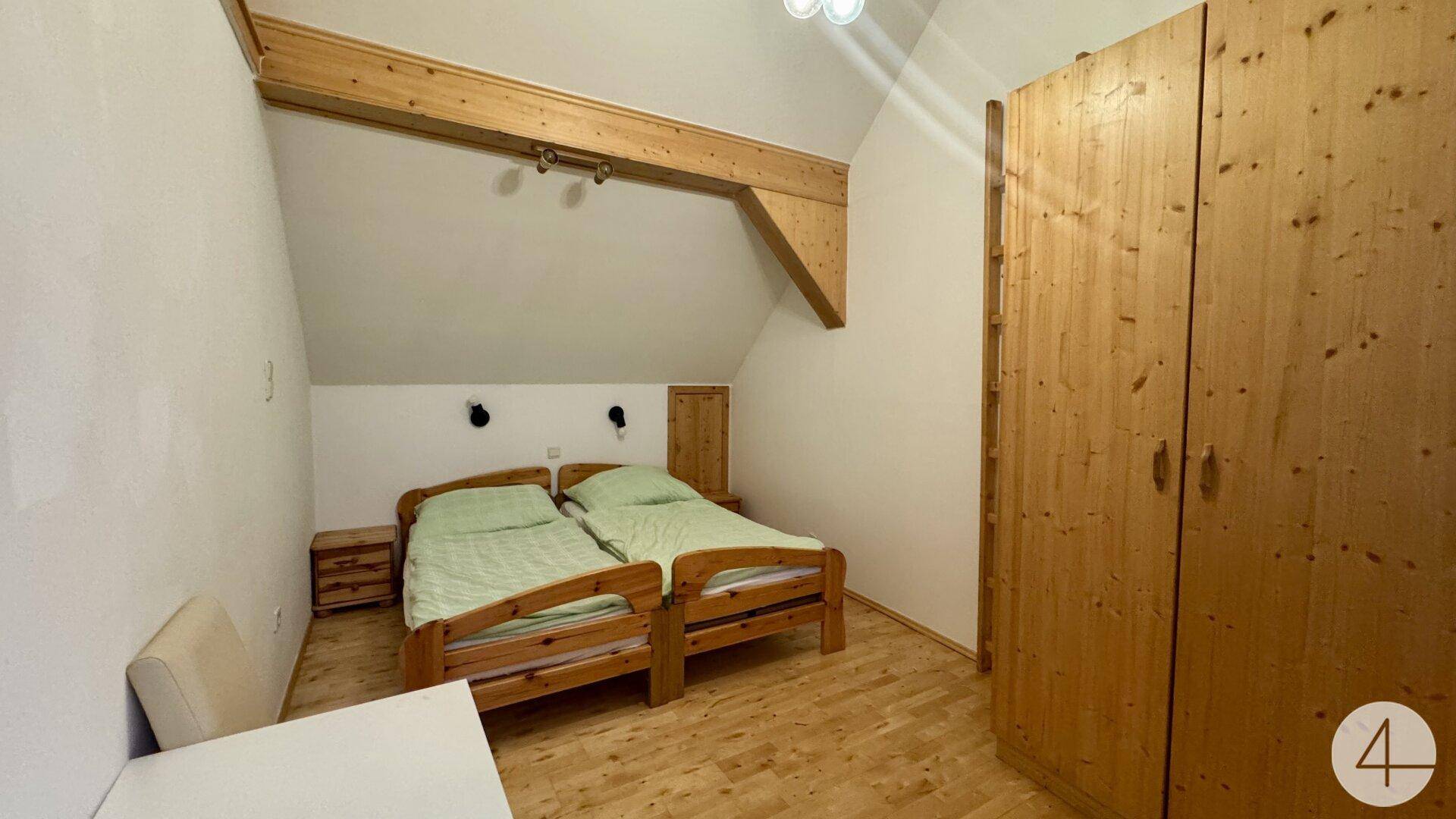 Schlafzimmer im Dachgeschoss mit zwei Einzelbetten, Holzschrank und sichtbaren Holzbalken an der Dachschräge.
