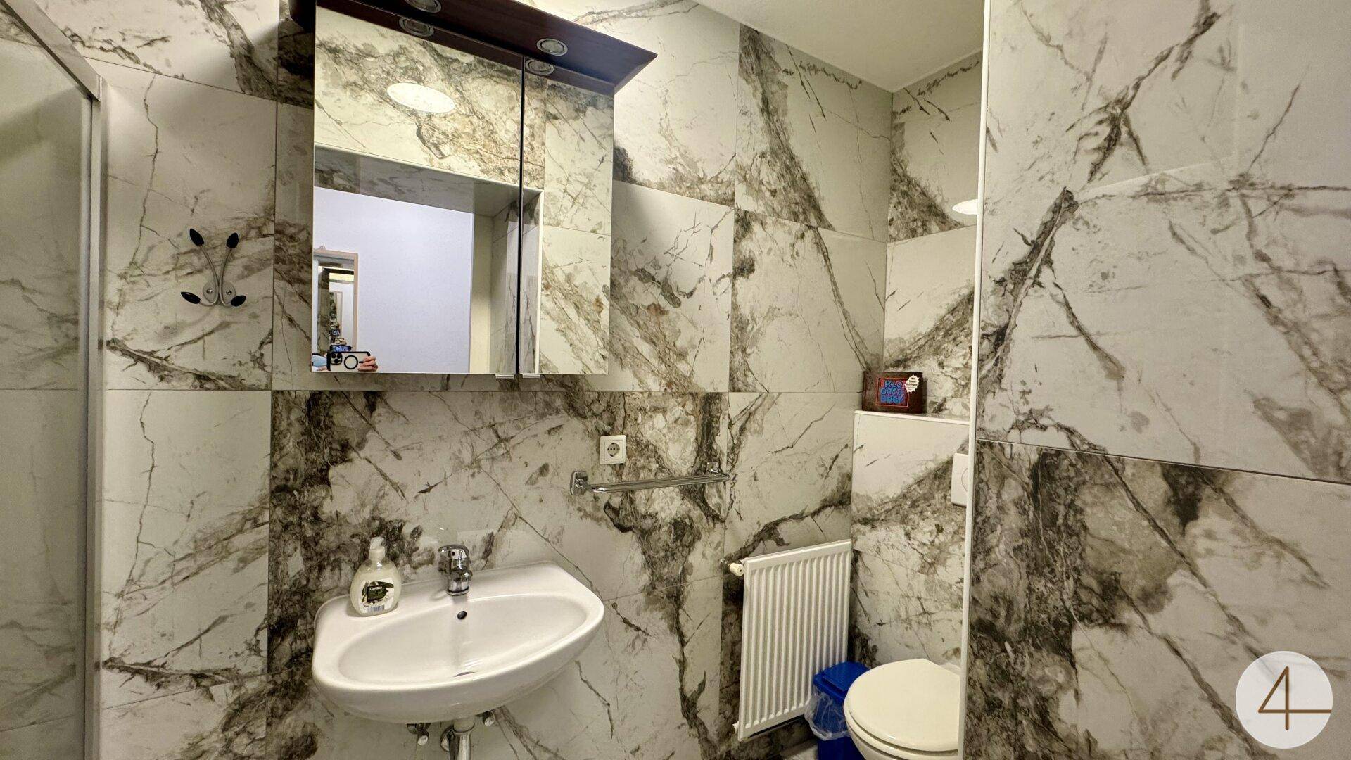 Badezimmer mit Marmor-Effekt-Fliesen, Waschbecken, Toilette und Spiegelschrank, inklusive Dusche.