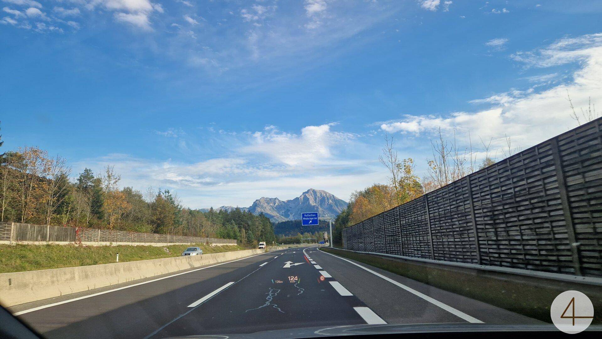 Blick von einer Autobahn auf ferne Berge, umgeben von Bäumen und einer Lärmschutzwand.