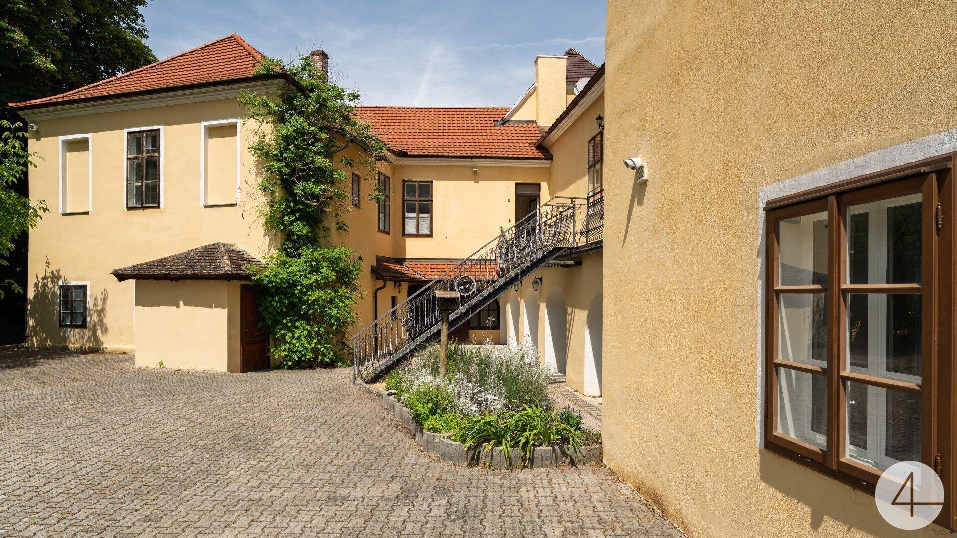 Innenhof eines historischen Gebäudes mit Außentreppe und gepflastertem Bereich, umgeben von Grün.