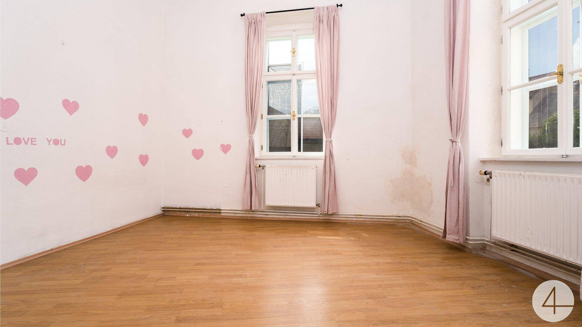 Helles Schlafzimmer mit Holzboden, zwei Fenstern und rosa Vorhängen, bereit zur Renovierung.