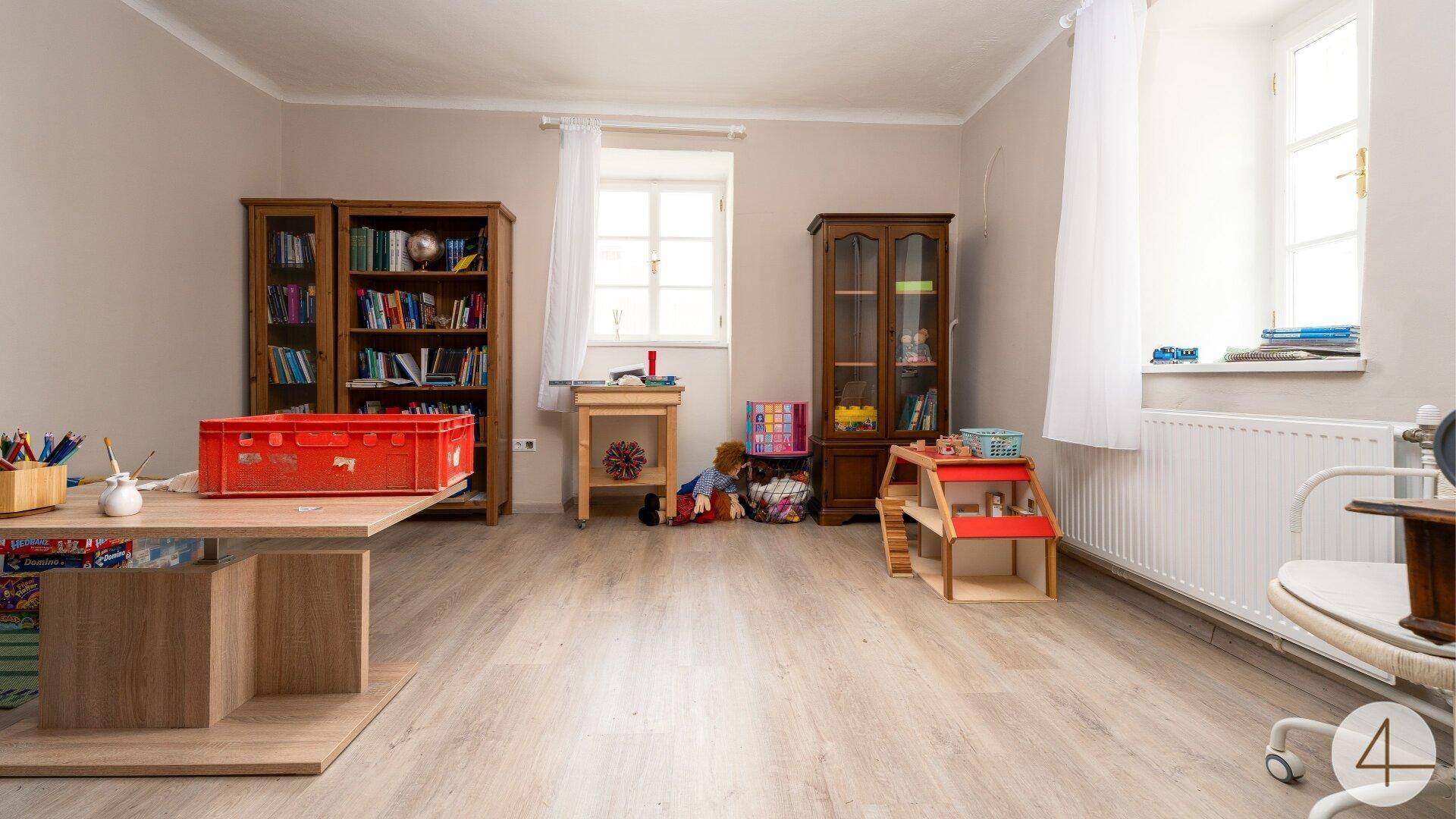 Heller Wohnraum mit Holzboden, zwei Fenstern und Bücherregalen, ideal für ein Kinderzimmer.