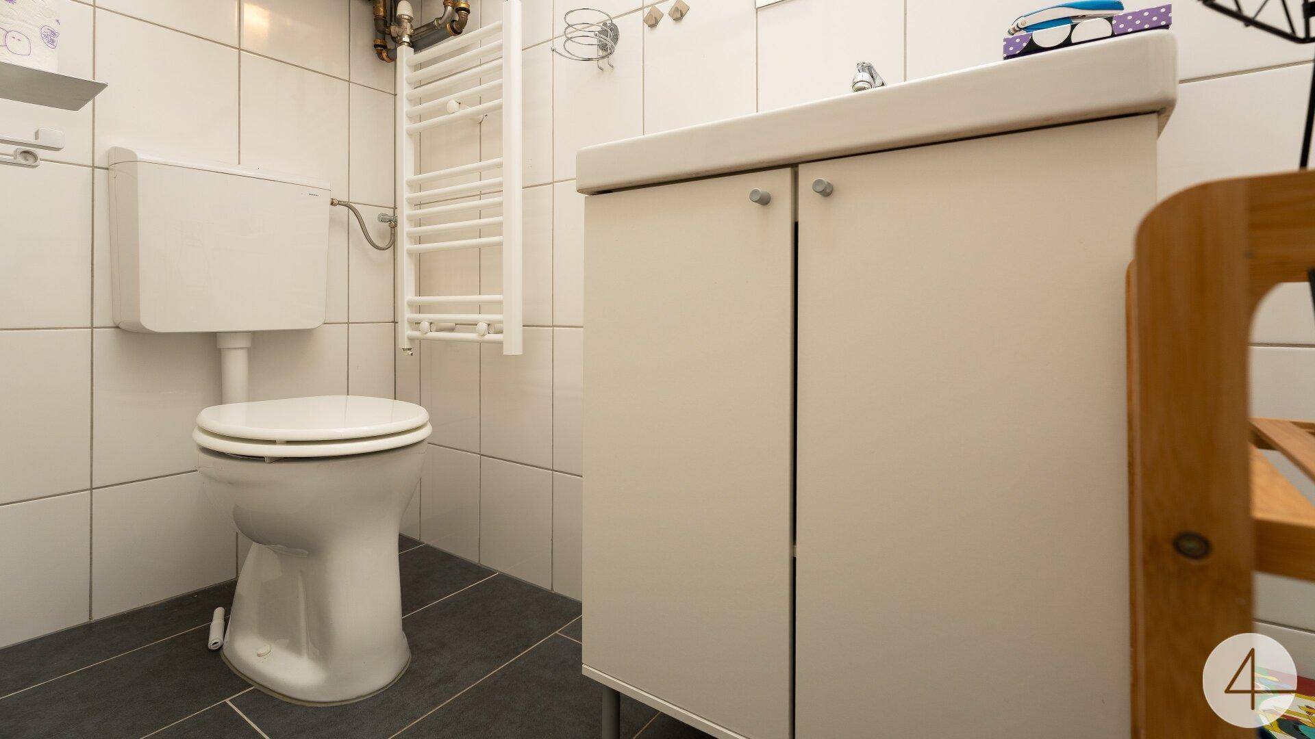 Kleines Badezimmer mit weißen Fliesen, Toilette und einem Waschbeckenunterschrank.
