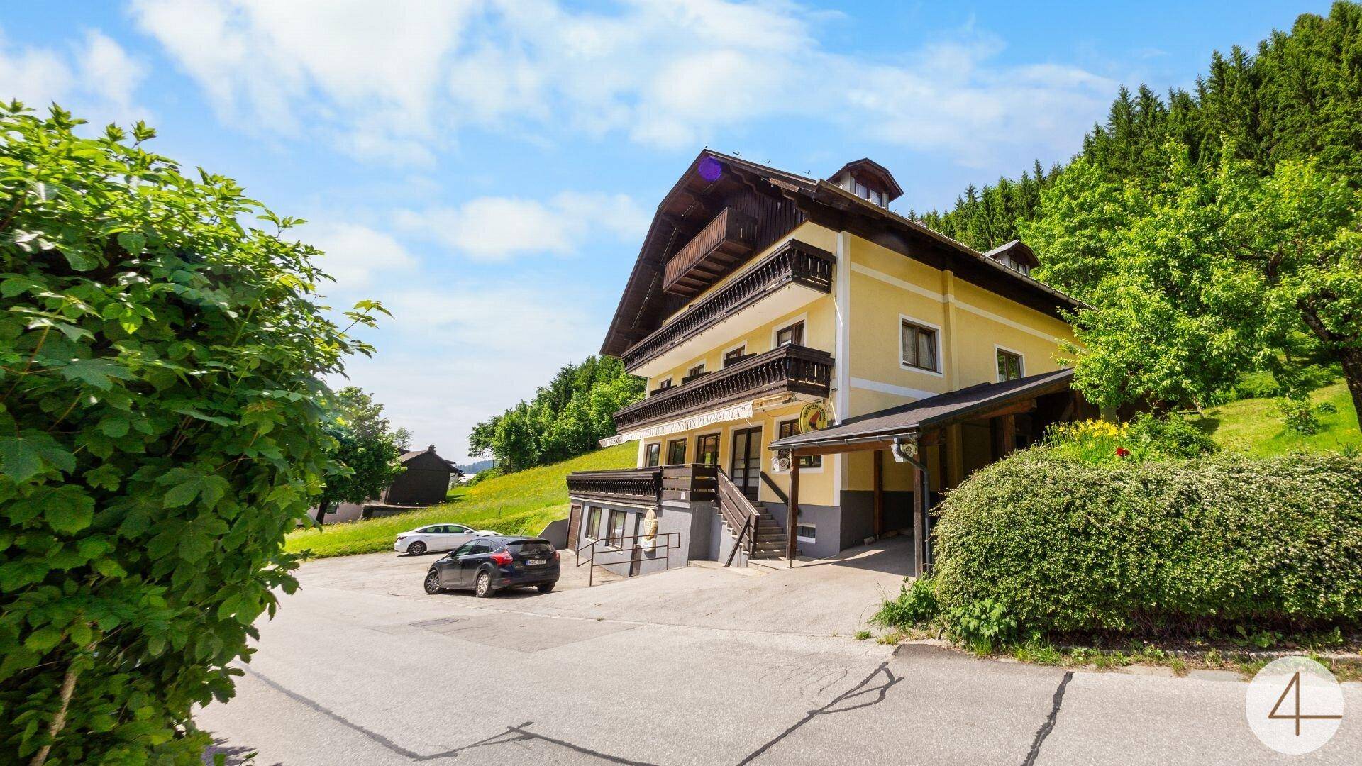 Ansicht des traditionellen Gebäudes mit Parkplätzen und üppiger grüner Umgebung unter blauem Himmel.