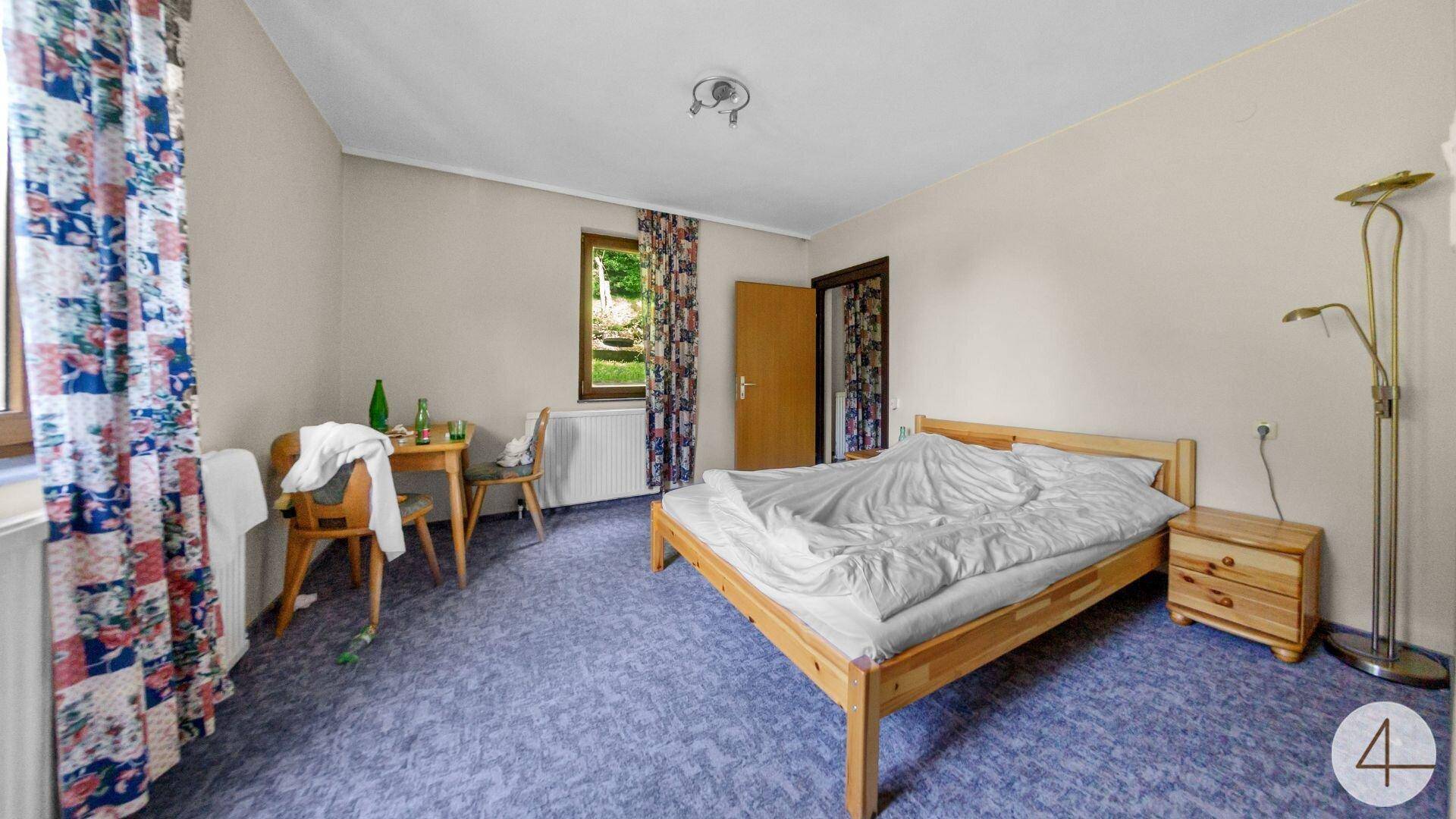 Schlafzimmer mit Einzelbett, Holzbettrahmen und blauem Teppichboden, Fenster mit Blick ins Grüne.