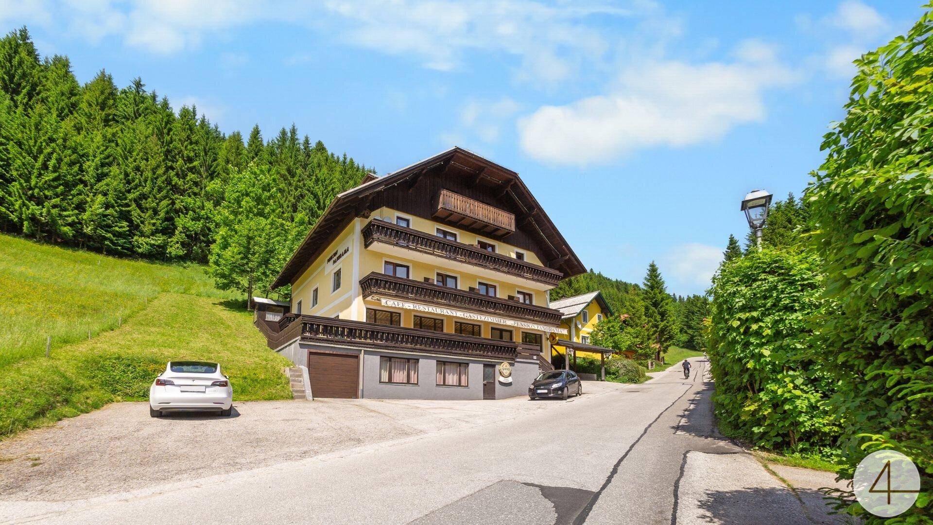 Traditionelles Gebäude in den Bergen, umgeben von grünen Wäldern und einer Zufahrtsstraße.