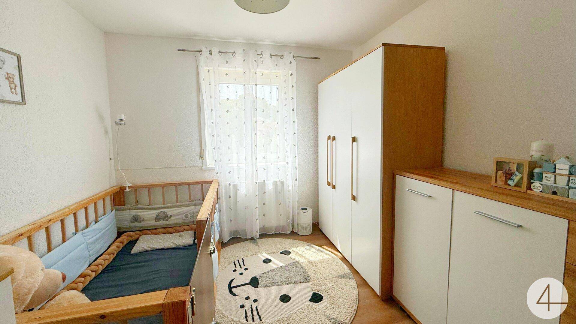 Kinderzimmer mit Babybett, weißem Kleiderschrank und viel Tageslicht durch das Fenster.