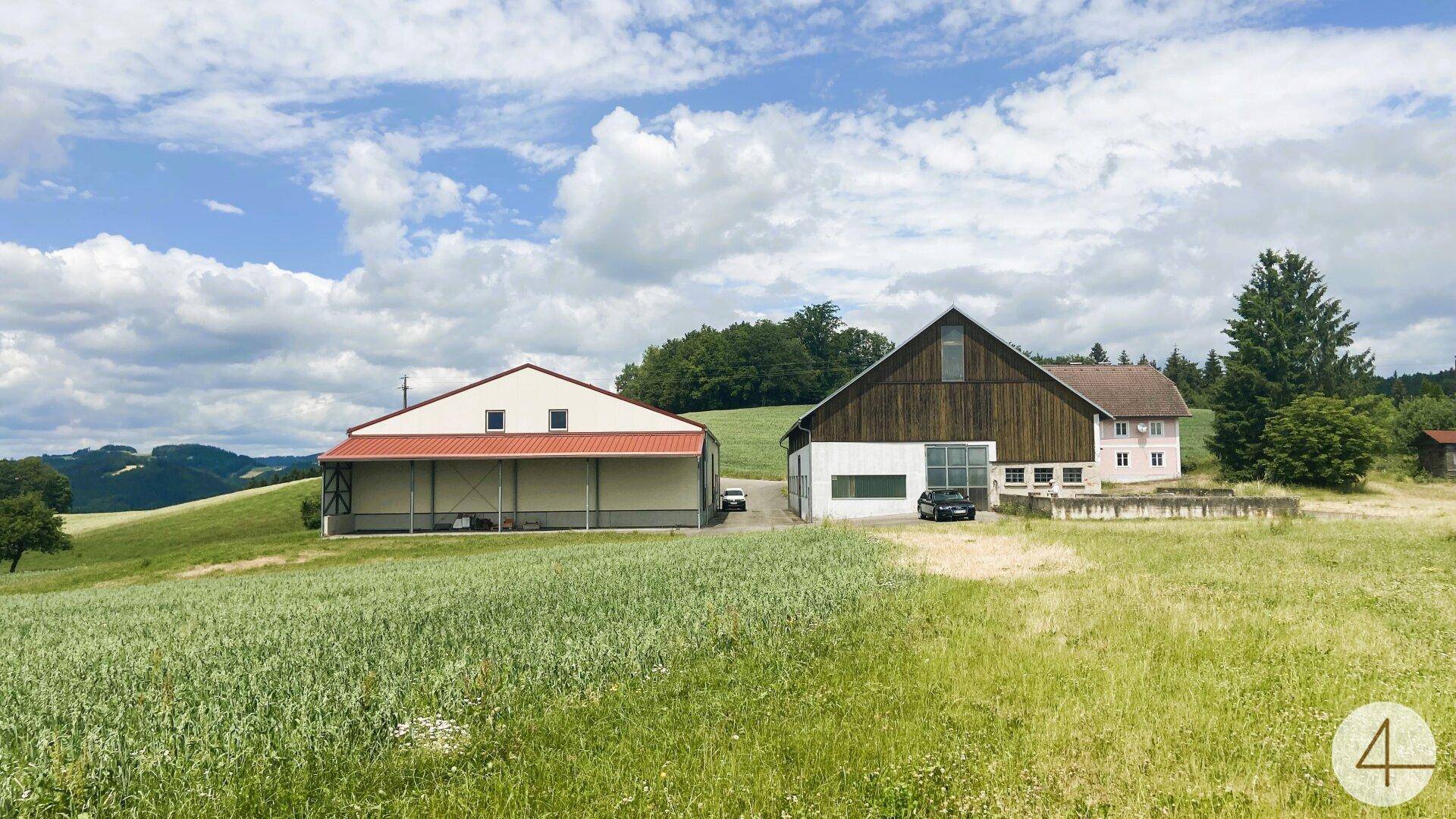 Weitläufige Ansicht von zwei Gebäuden in ländlicher Umgebung mit Feldern und Hügeln unter blauem Himmel.