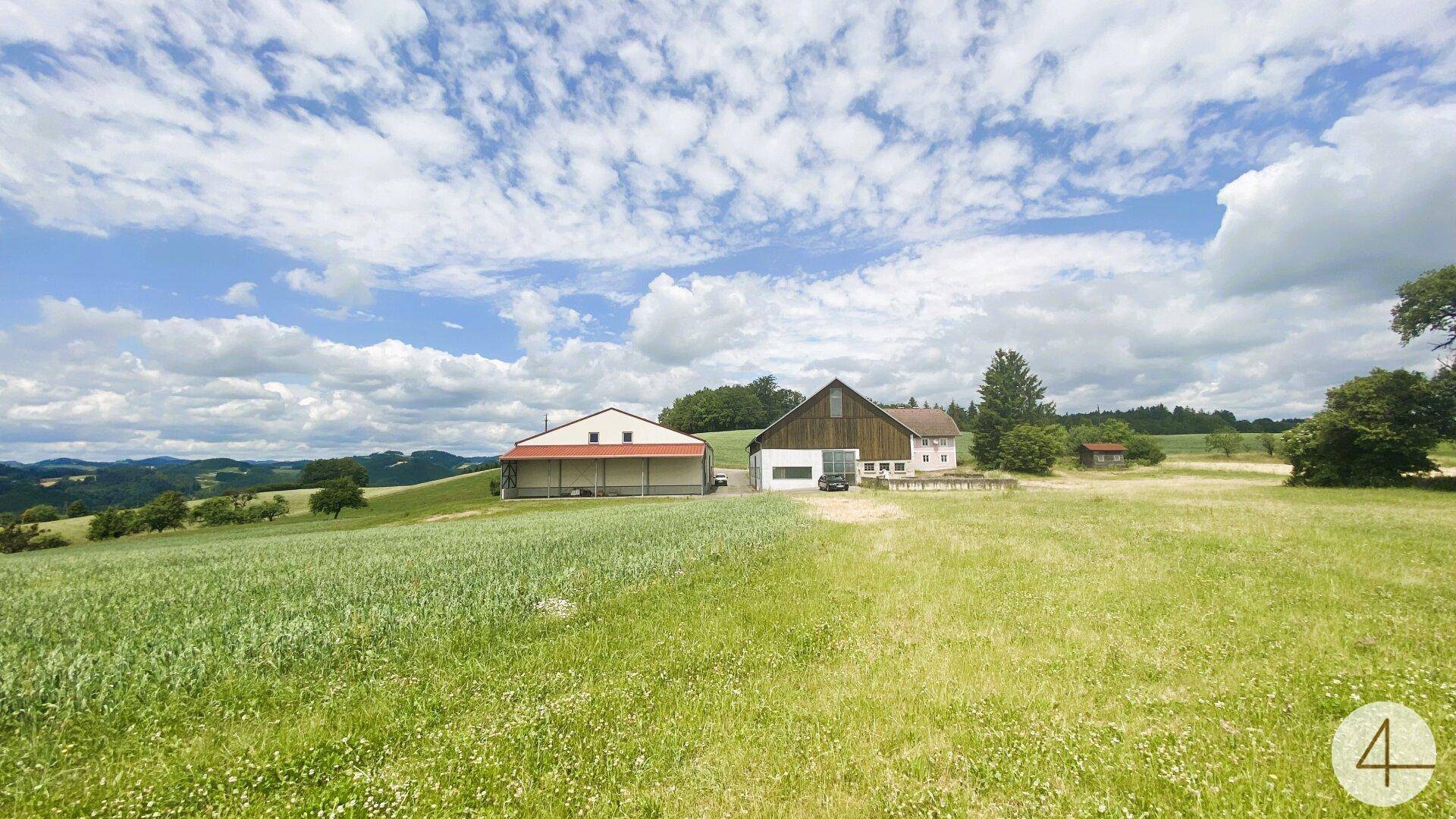 Panoramablick auf zwei Gebäude in einer weiten, grünen Landschaft mit Feldern und bewaldeten Hügeln.