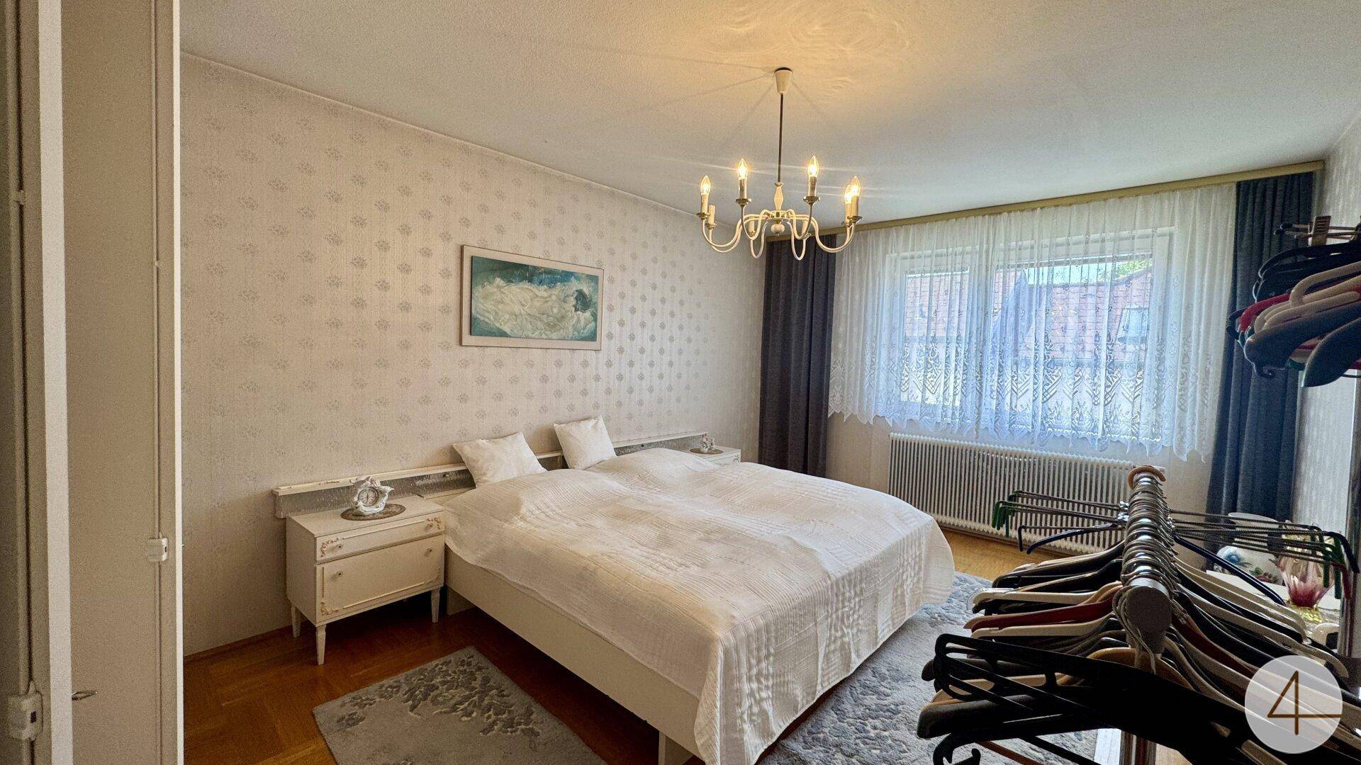 Schlafzimmer mit Doppelbett, Nachttischen und Kronleuchter, hell durch großes Fenster.