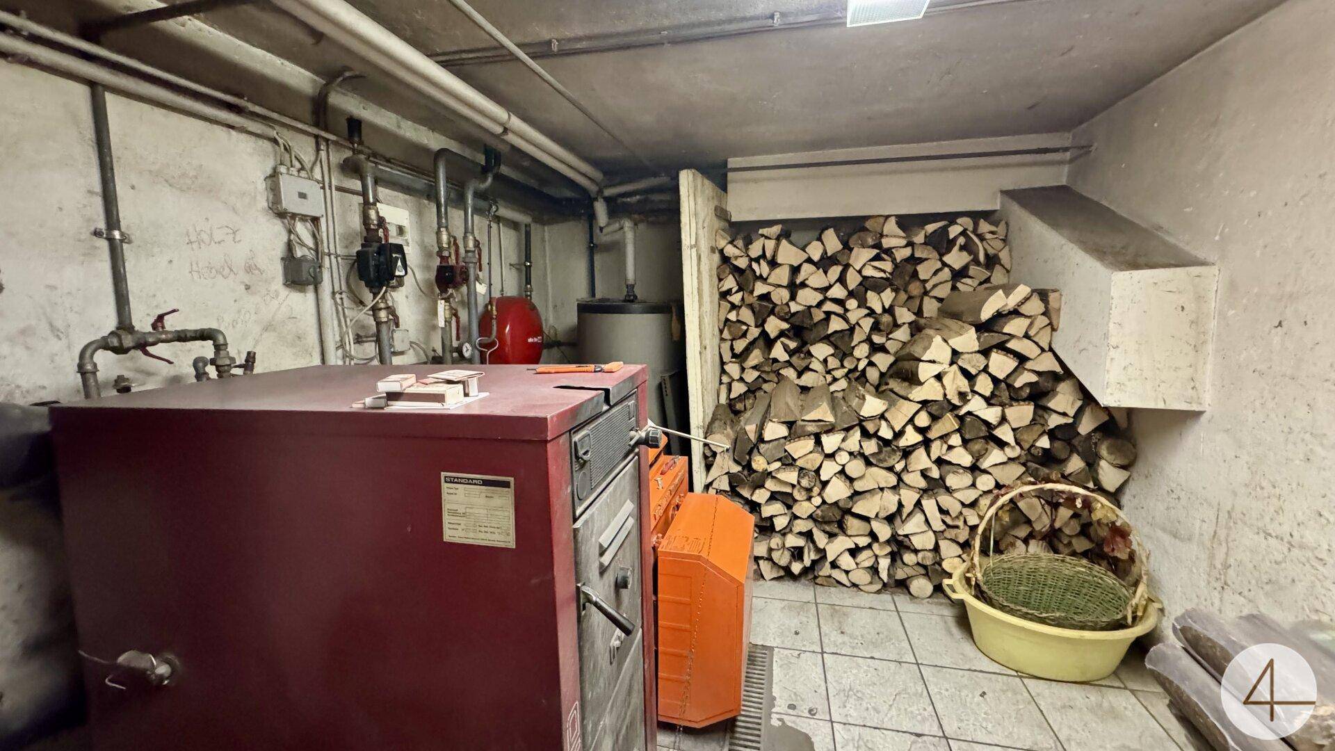 Kellerraum mit Heizungsanlage, Rohrleitungen und einem großen Stapel Brennholz.