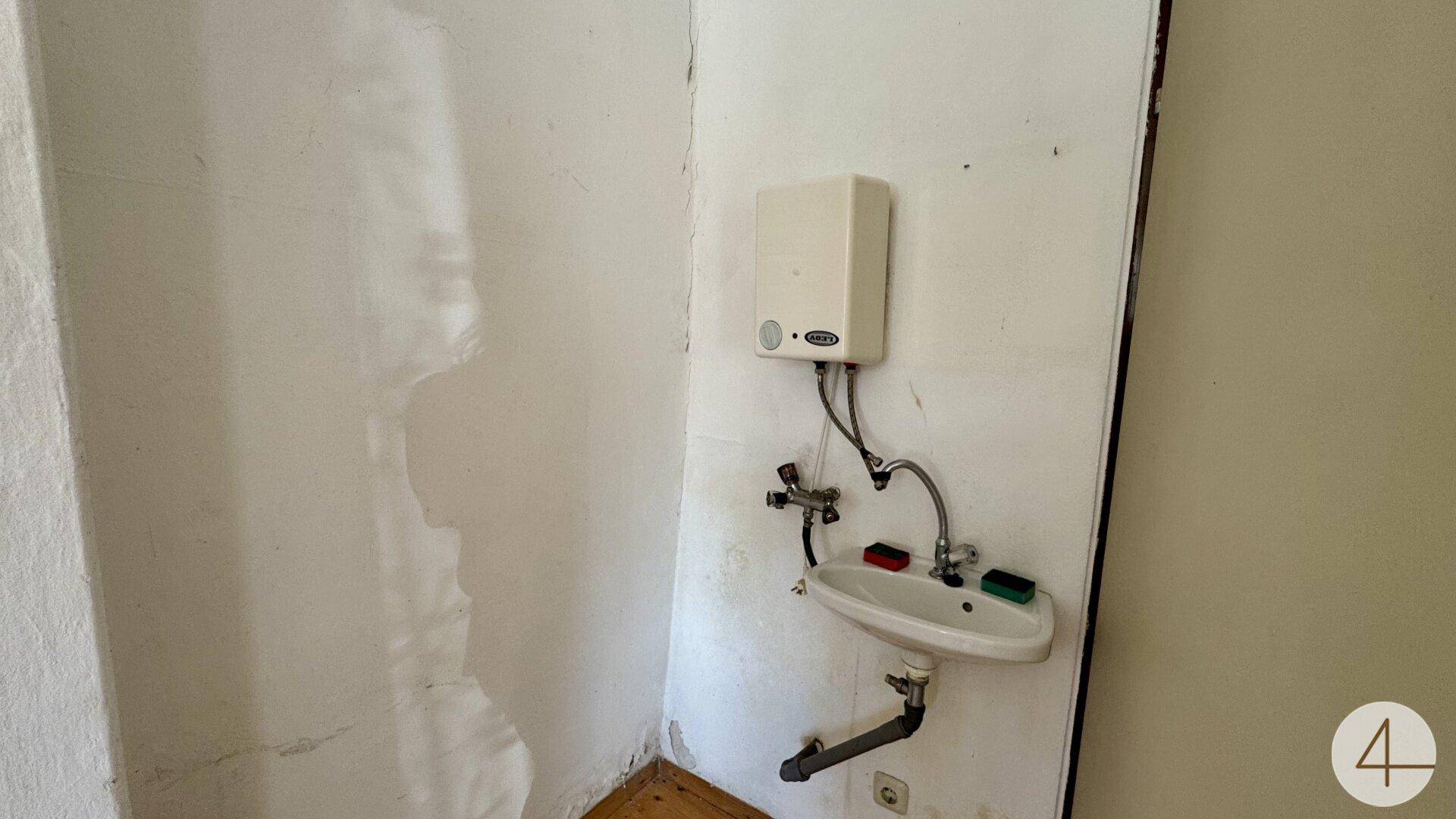 Einfacher Waschbereich mit kleinem Waschbecken und Warmwasserbereiter an weißer Wand.